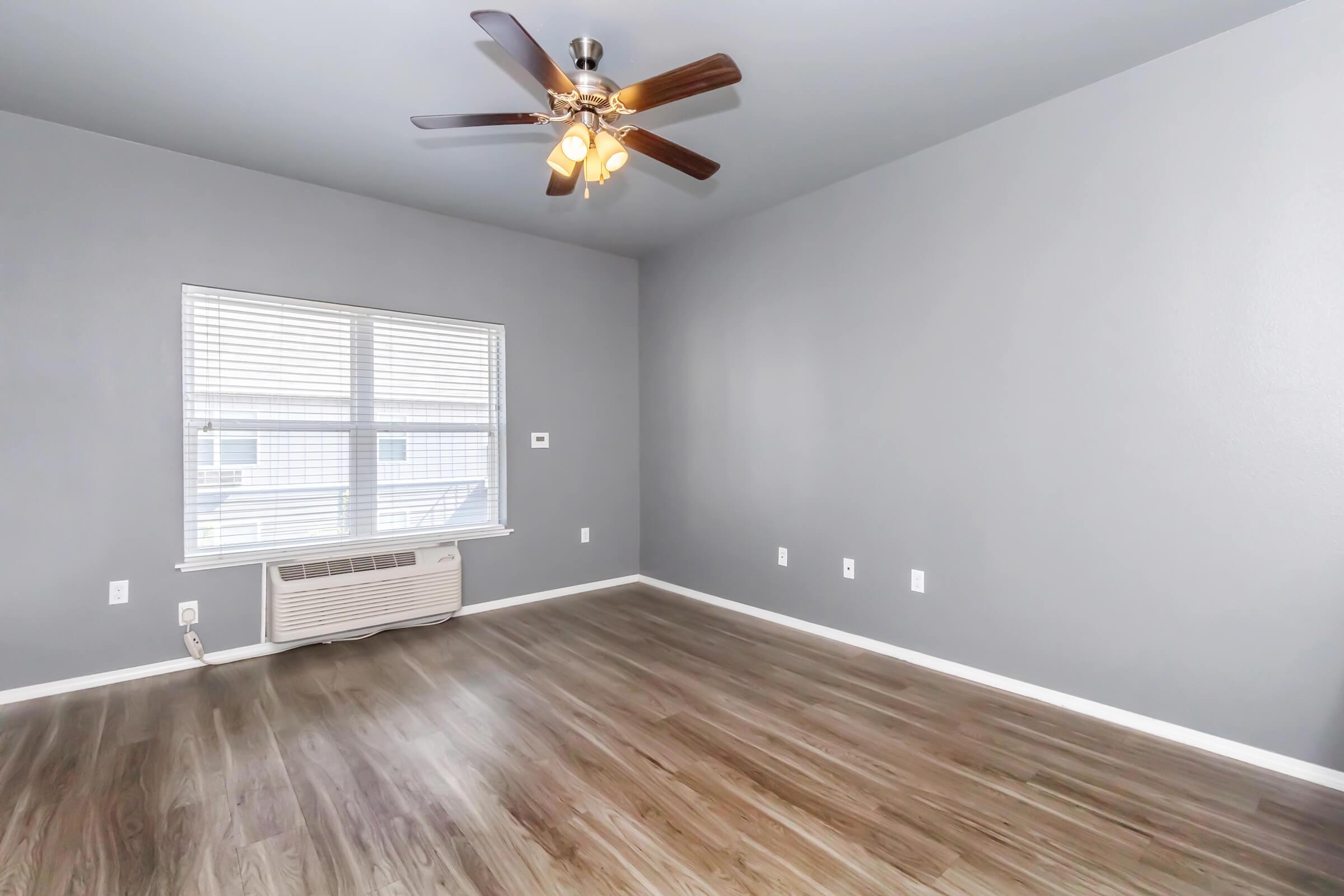 A spacious room featuring grey walls and a ceiling fan with lights. The flooring is wood-like laminate, and there is a window with blinds allowing natural light. An air conditioning unit is visible on the wall, and the overall space is empty, ready for decoration.