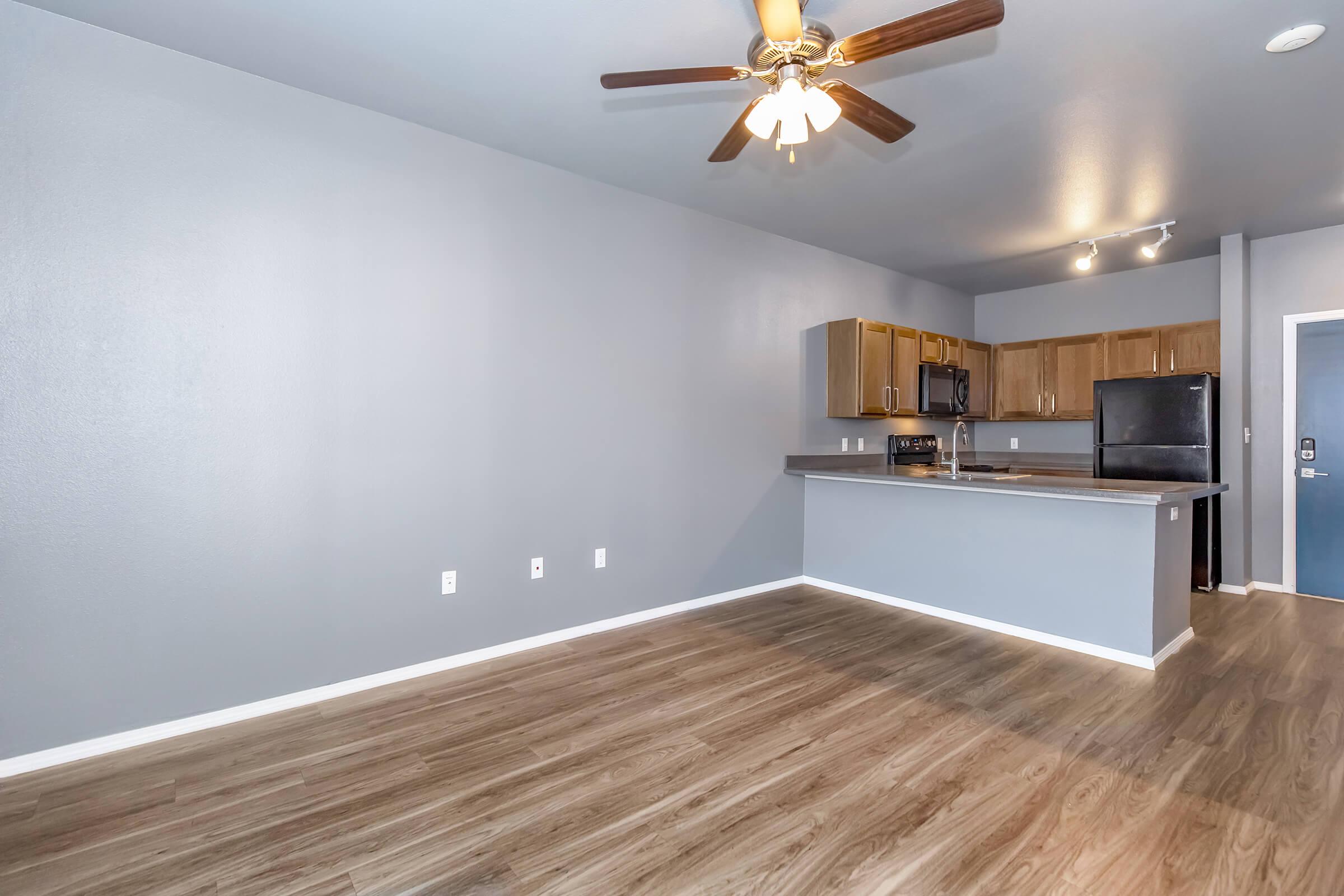 A spacious, modern living area featuring light gray walls and a wooden floor. An open kitchen with wooden cabinets, a black refrigerator, and a microwave. Ceiling fan with light fixture. The layout offers a clean, airy atmosphere, perfect for comfortable living.