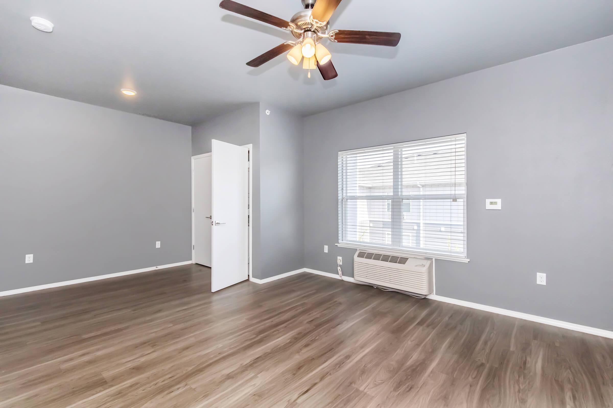 A spacious room with gray walls and a ceiling fan. There is a window with blinds allowing natural light, a door leading to another space, and an air conditioning unit on the wall. The floor is made of light-colored wood laminate, creating a modern and inviting atmosphere.