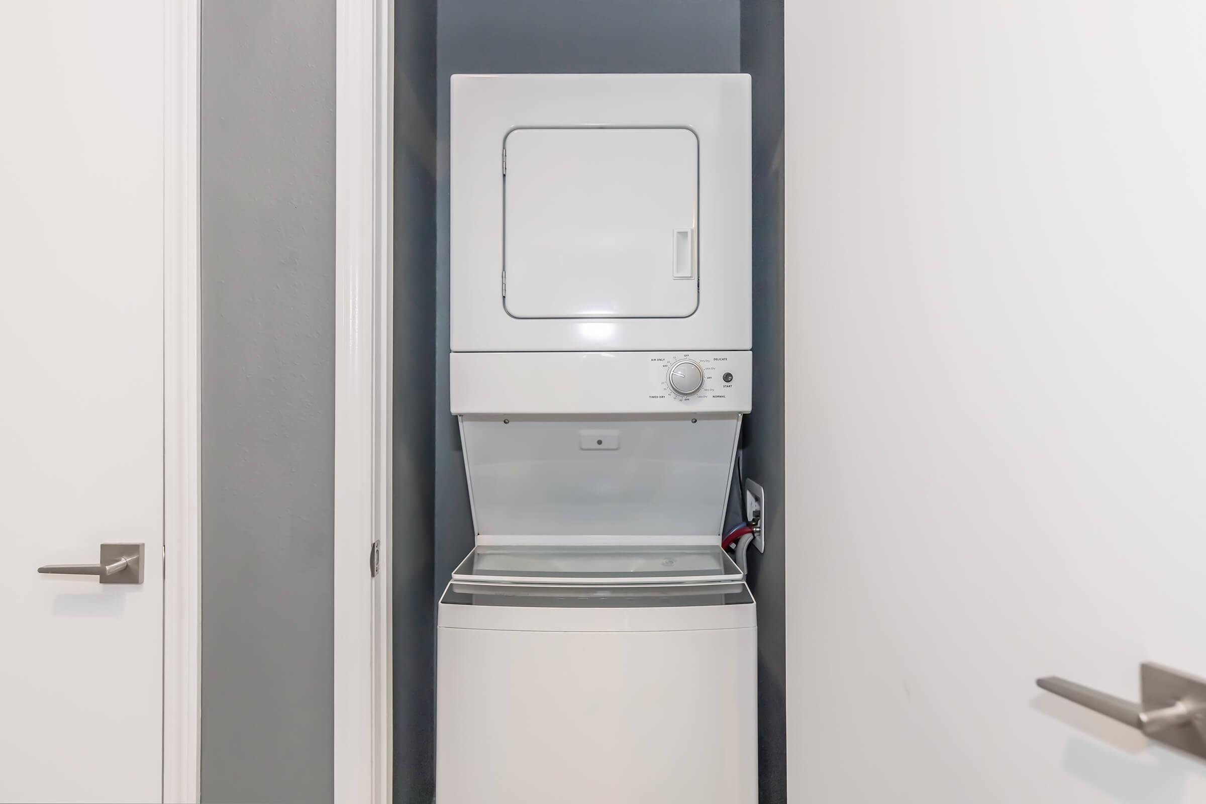 A stacked washer and dryer unit located in a narrow laundry space, with a light gray wall in the background and white doors flanking the unit. The appliances are modern and compact, designed for efficient use of space.