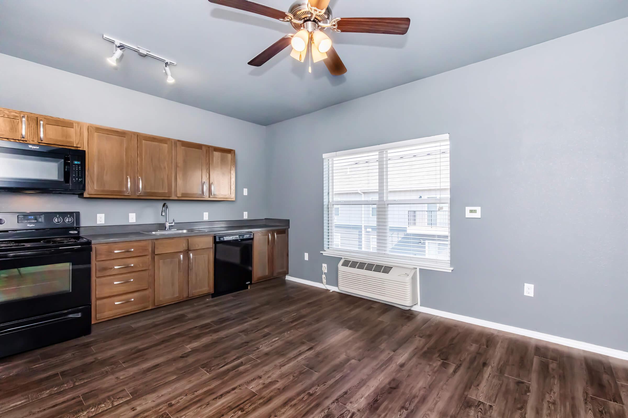 A modern kitchen featuring wooden cabinets, black appliances including an oven and dishwasher, and a ceiling fan. The walls are painted a soft gray, and there’s a large window providing natural light. The flooring is dark wood, and there is an air conditioning unit beneath the window.