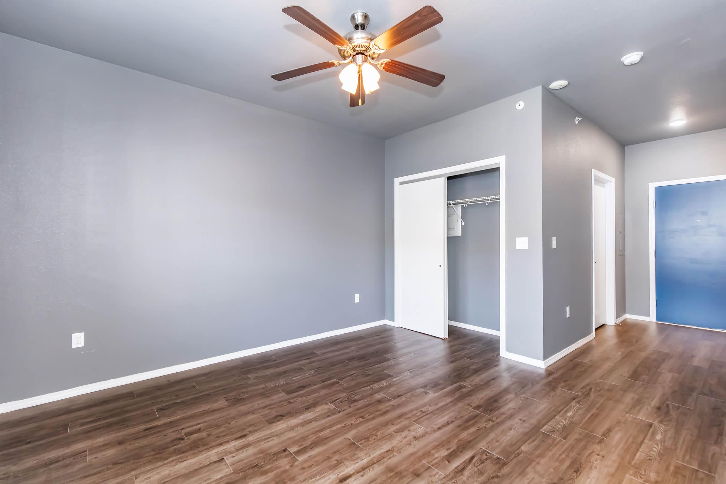 A well-lit room featuring a ceiling fan, gray walls, and wooden flooring. There's a closet with a sliding door on the left, and a doorway leading to another room on the right. The overall atmosphere is spacious and modern, with a clean and inviting aesthetic.