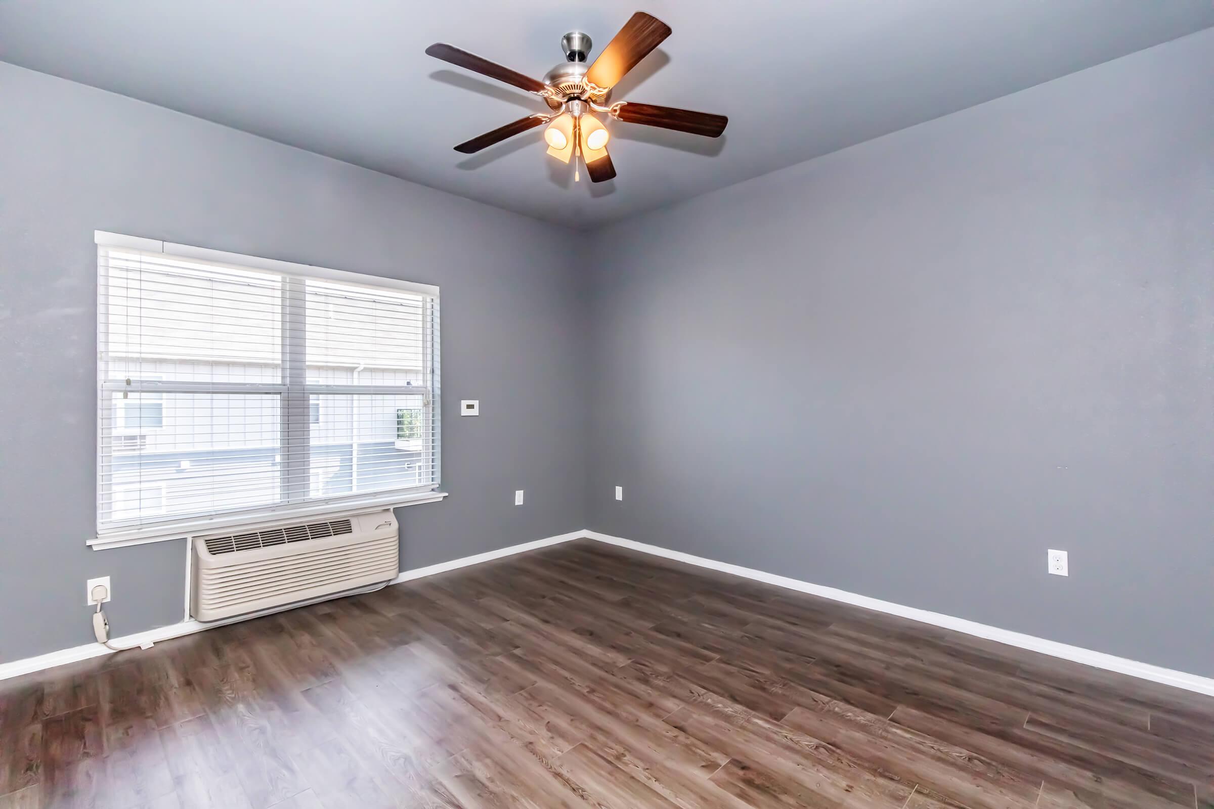 A spacious room with gray walls and a ceiling fan. The floor is made of wood laminate, and there’s a large window allowing natural light. An air conditioning unit is positioned below the window, creating a modern and comfortable atmosphere.