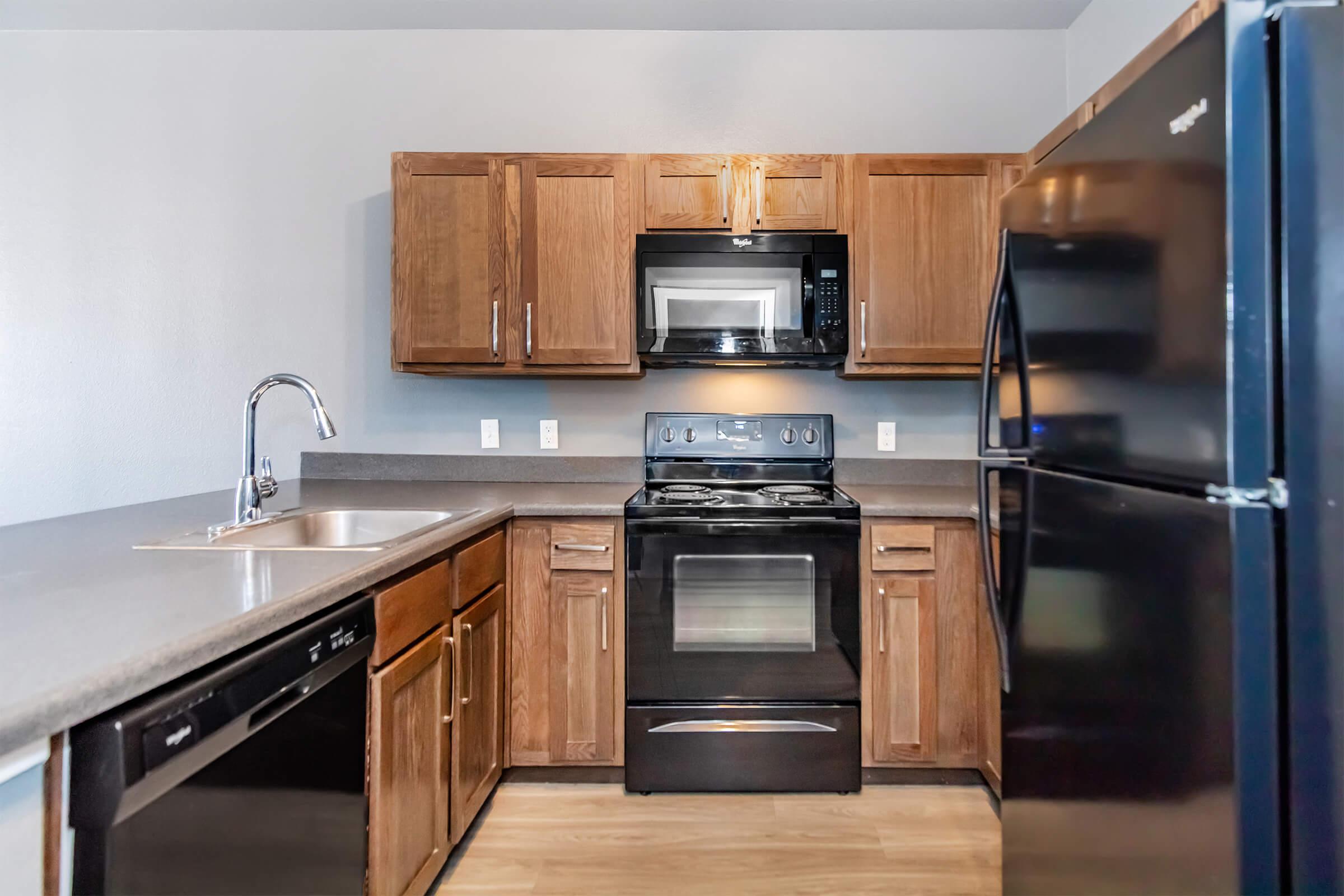 A modern kitchen featuring wooden cabinets, a stainless steel sink, a black oven, a black microwave above it, and a black refrigerator. The countertop is gray, and the walls are painted light blue, creating a clean and contemporary look.
