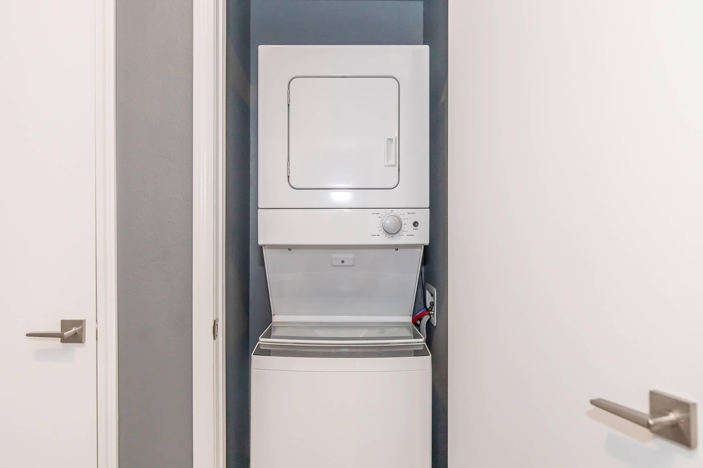 A stacked washer and dryer unit tucked into a narrow space between two white doors, with a gray wall in the background. The appliance has a control panel and is positioned above a washer, showcasing an efficient use of space in a laundry area.