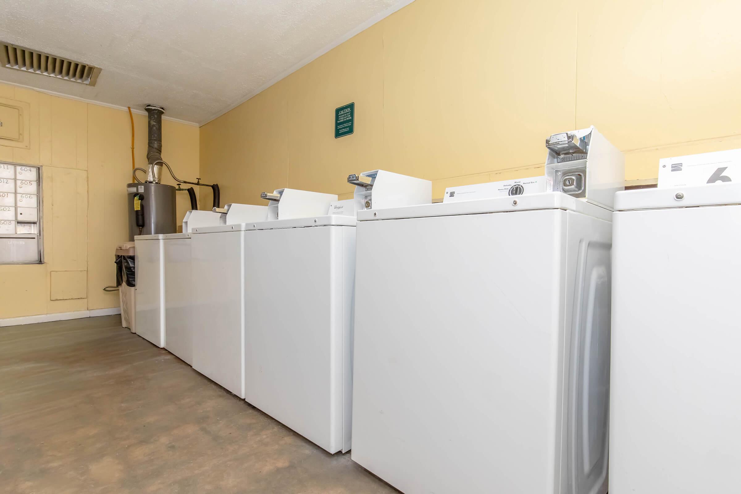 A row of white laundry machines lined up against a yellow wall in a laundry room, with a ventilation duct visible on the ceiling and a wall-mounted sign above the machines. The floor is concrete, and the setting appears clean and organized.