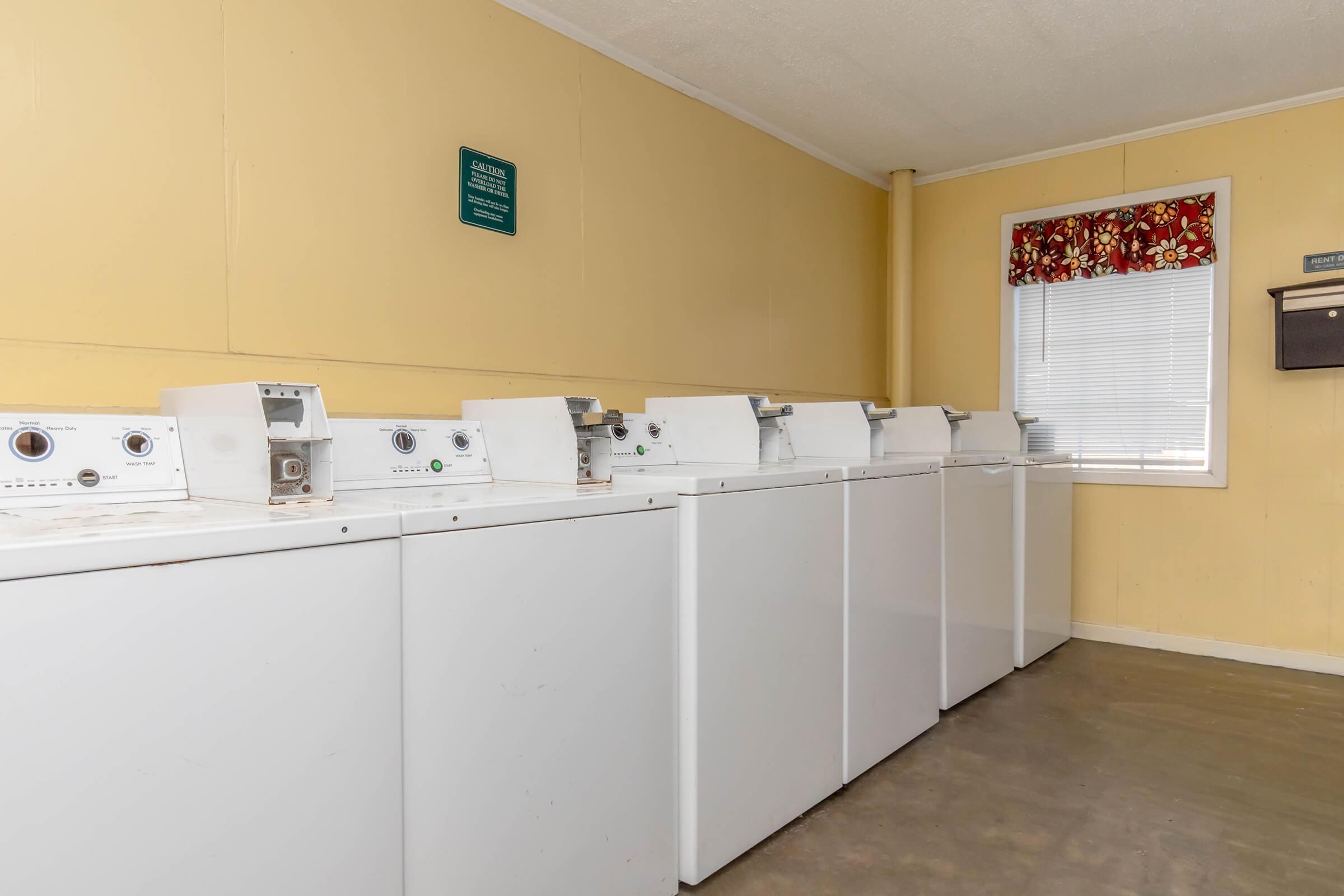 A row of white washing machines lined up against a yellow wall in a laundry room. There is a window with a floral curtain and a wall-mounted plaque or sign above the machines. The floor is a bare concrete surface, and the atmosphere is clean and utilitarian.
