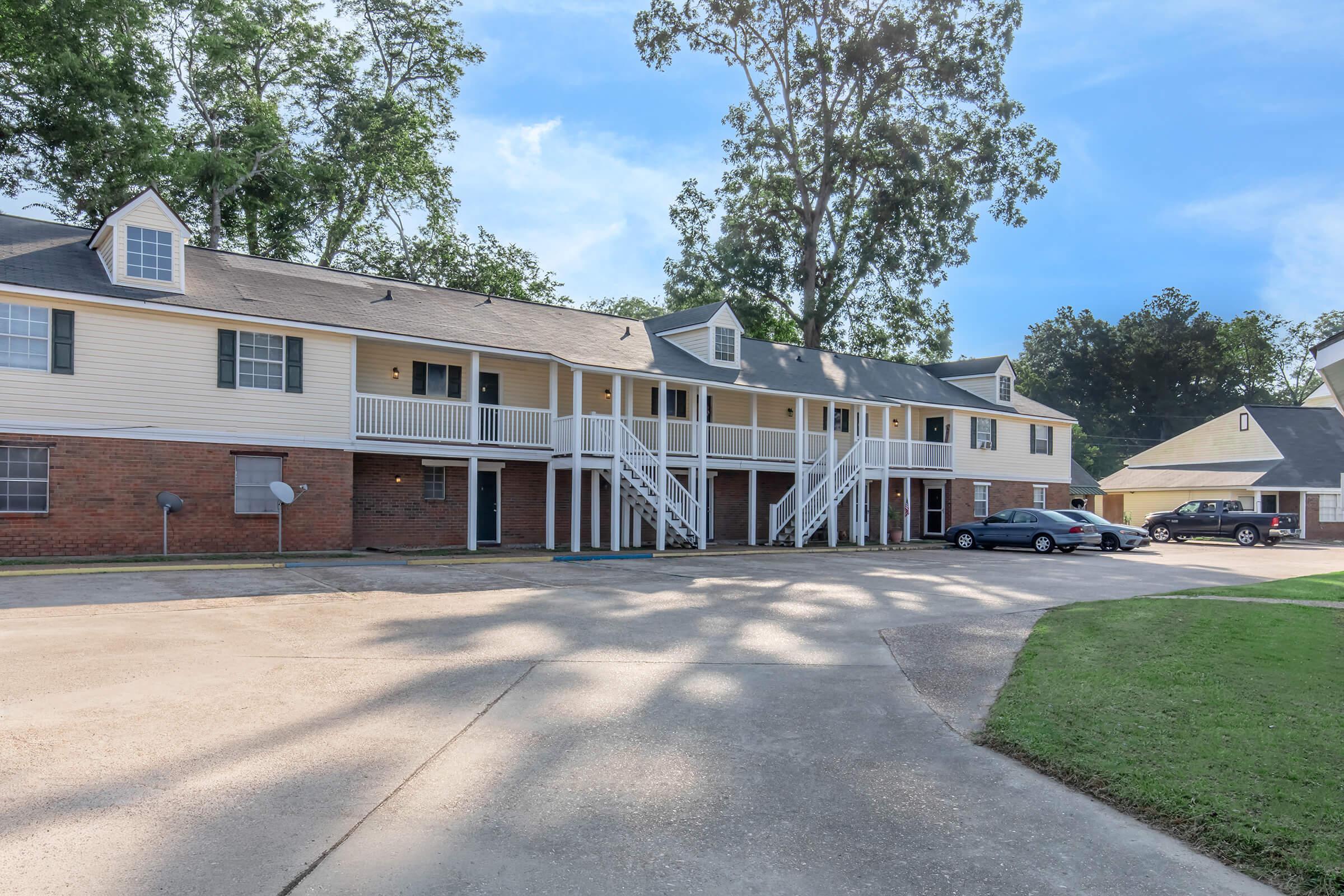 A low-rise apartment building featuring a mix of brick and cream-colored siding. The building has multiple entrances with white railings, shaded by trees. An asphalt driveway runs in front, with grassy areas on the sides. Clear blue sky and soft shadows create a peaceful atmosphere.