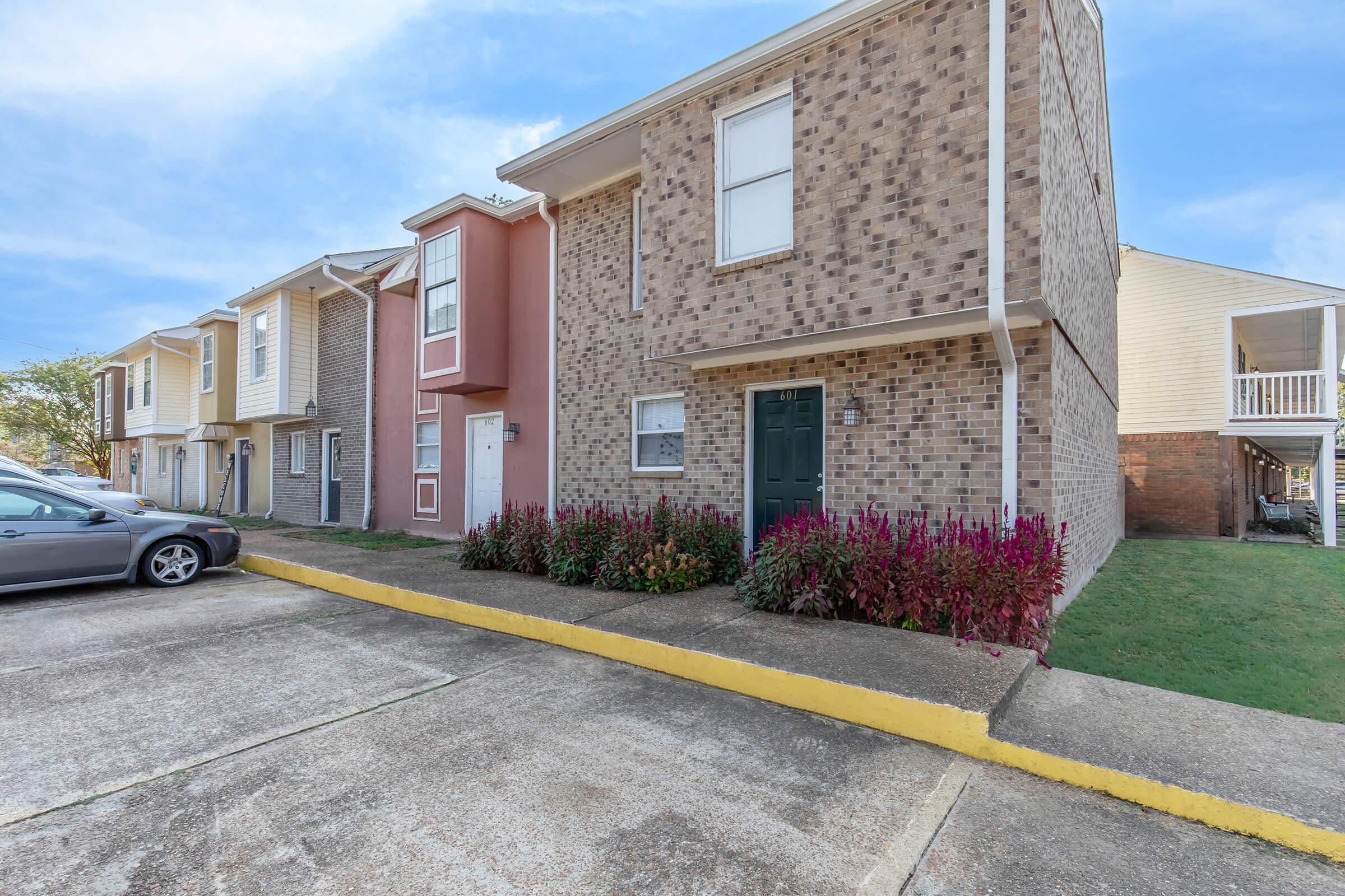 A row of townhouse-style apartments with varying brick and siding colors. The foreground features a flowerbed with colorful plants, and a parked car is visible nearby. The scene is set against a clear blue sky, providing a bright and inviting atmosphere.