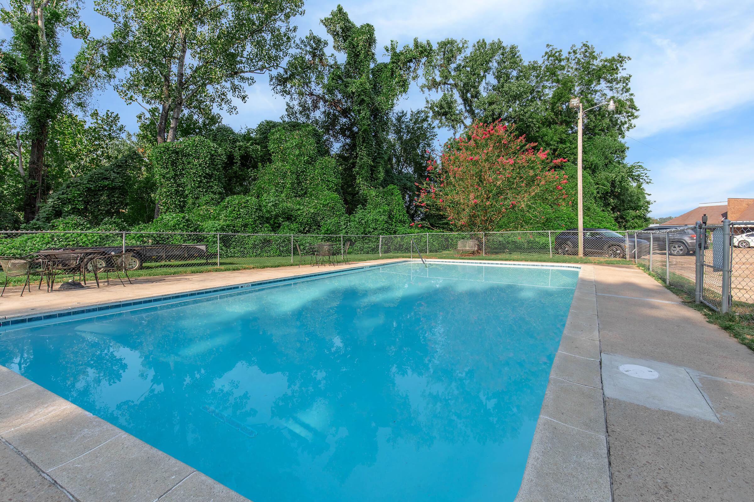 A clear blue swimming pool surrounded by a fenced area. Lush green trees and vegetation are visible in the background, along with a vibrant flowering plant. The pool area includes lounge chairs and a pathway, creating a serene outdoor environment.