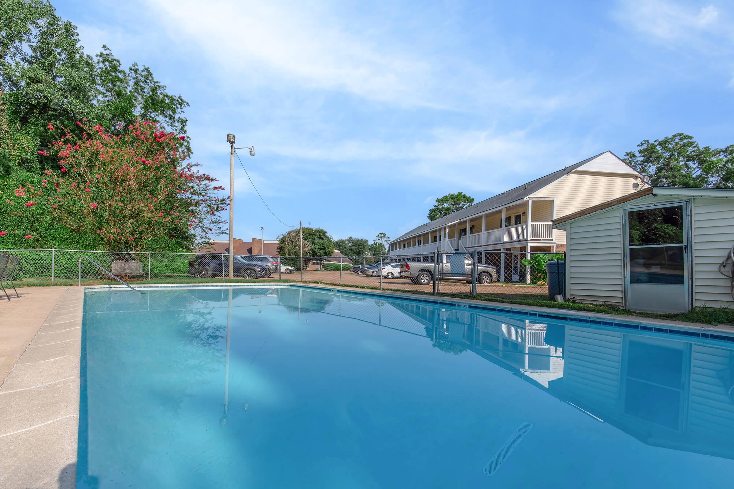 View of a clear blue swimming pool with a surrounding concrete deck. In the background, there are two multi-story buildings, greenery with flowering plants on one side, and parked cars visible nearby. The sky is bright with a few clouds, creating a calm and inviting atmosphere.