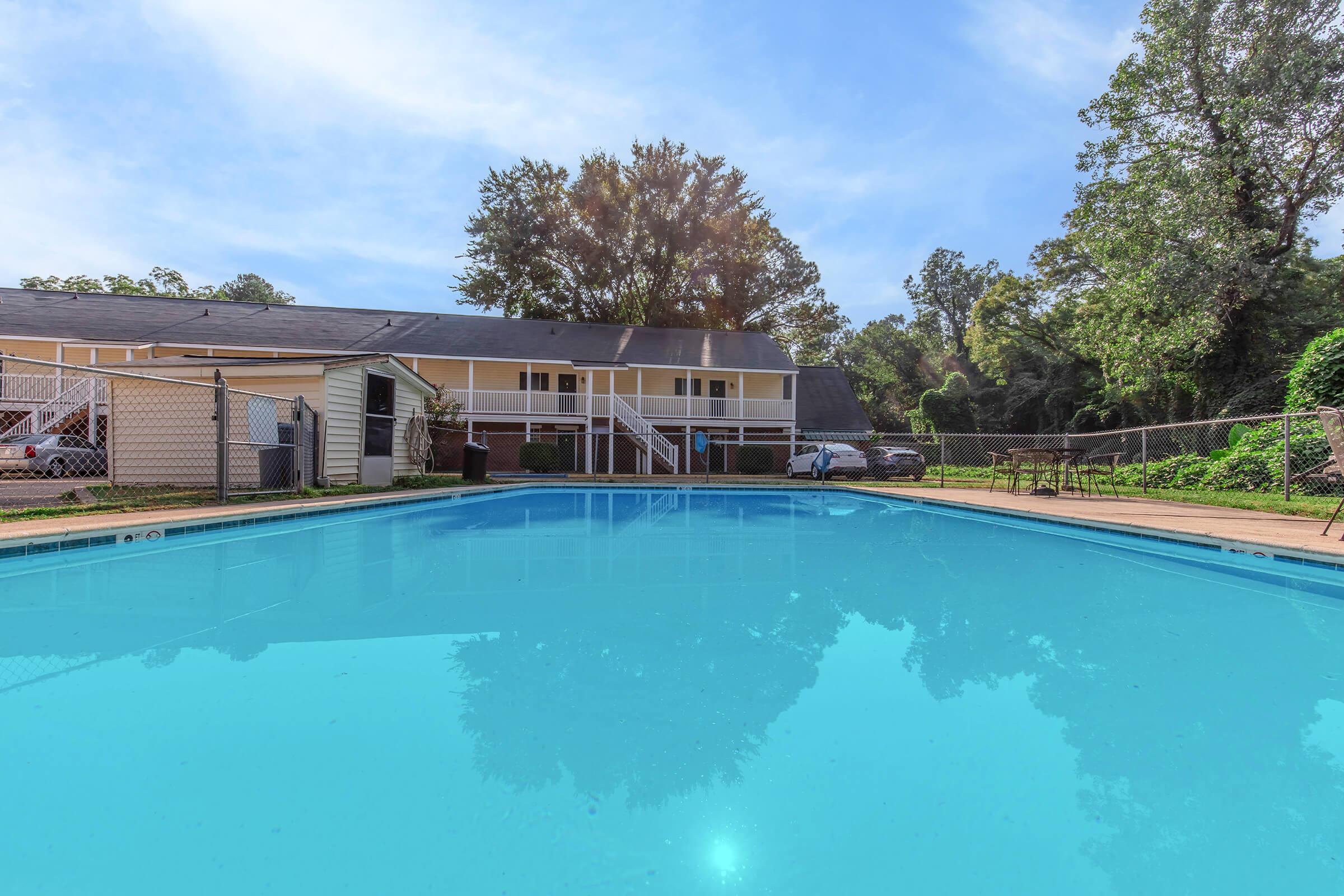 A clear swimming pool reflecting the sky in the foreground, with a fenced area surrounding it. In the background, a two-story building with multiple balconies is visible, along with some trees and vehicles. The scene is bright and inviting, showcasing a peaceful outdoor setting.
