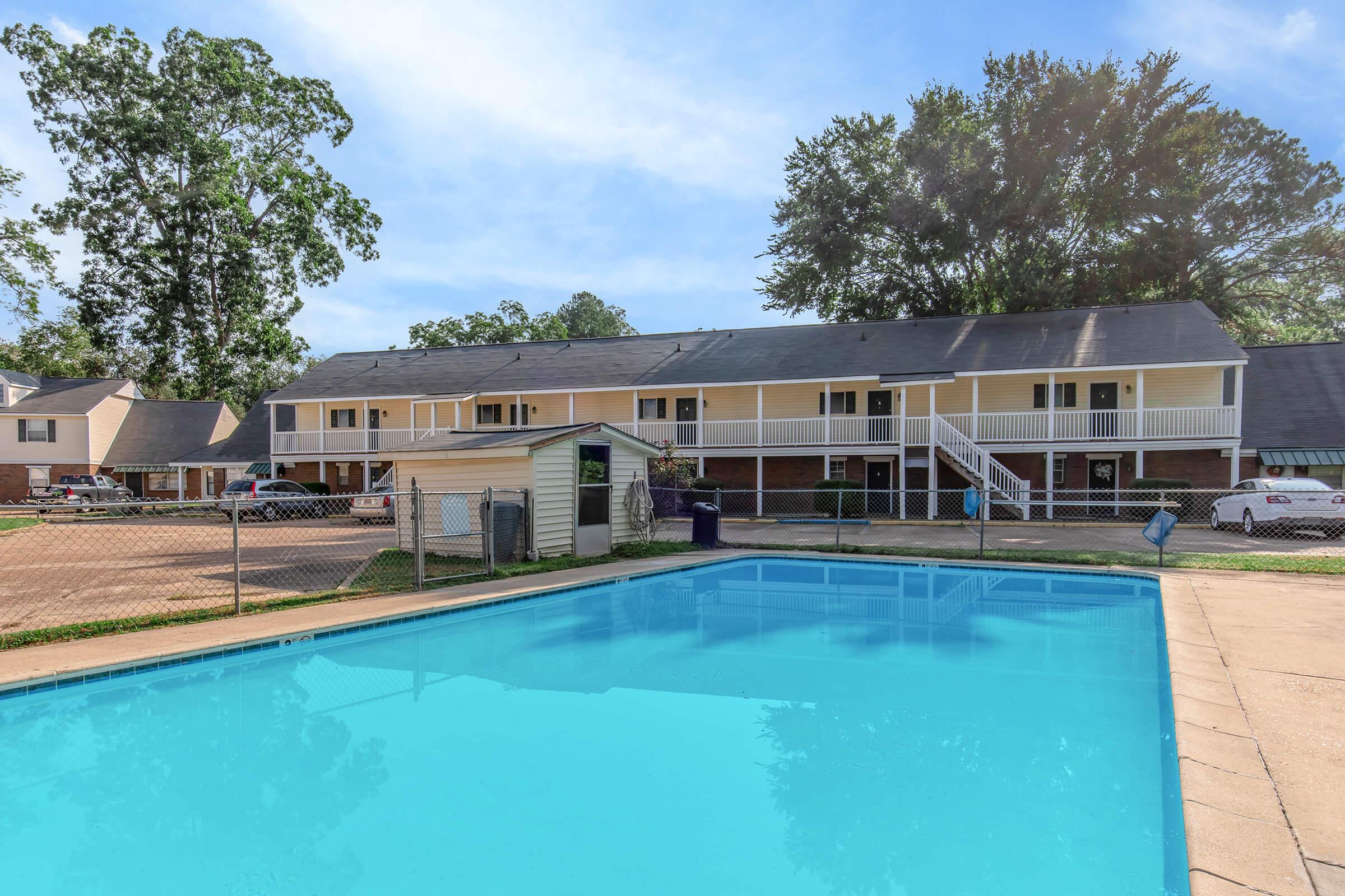 A view of a well-maintained outdoor swimming pool in front of a two-story apartment building. The building features a porch with white railings and is surrounded by trees, creating a peaceful environment. Cars are parked nearby, and there's a small shed by the pool area.