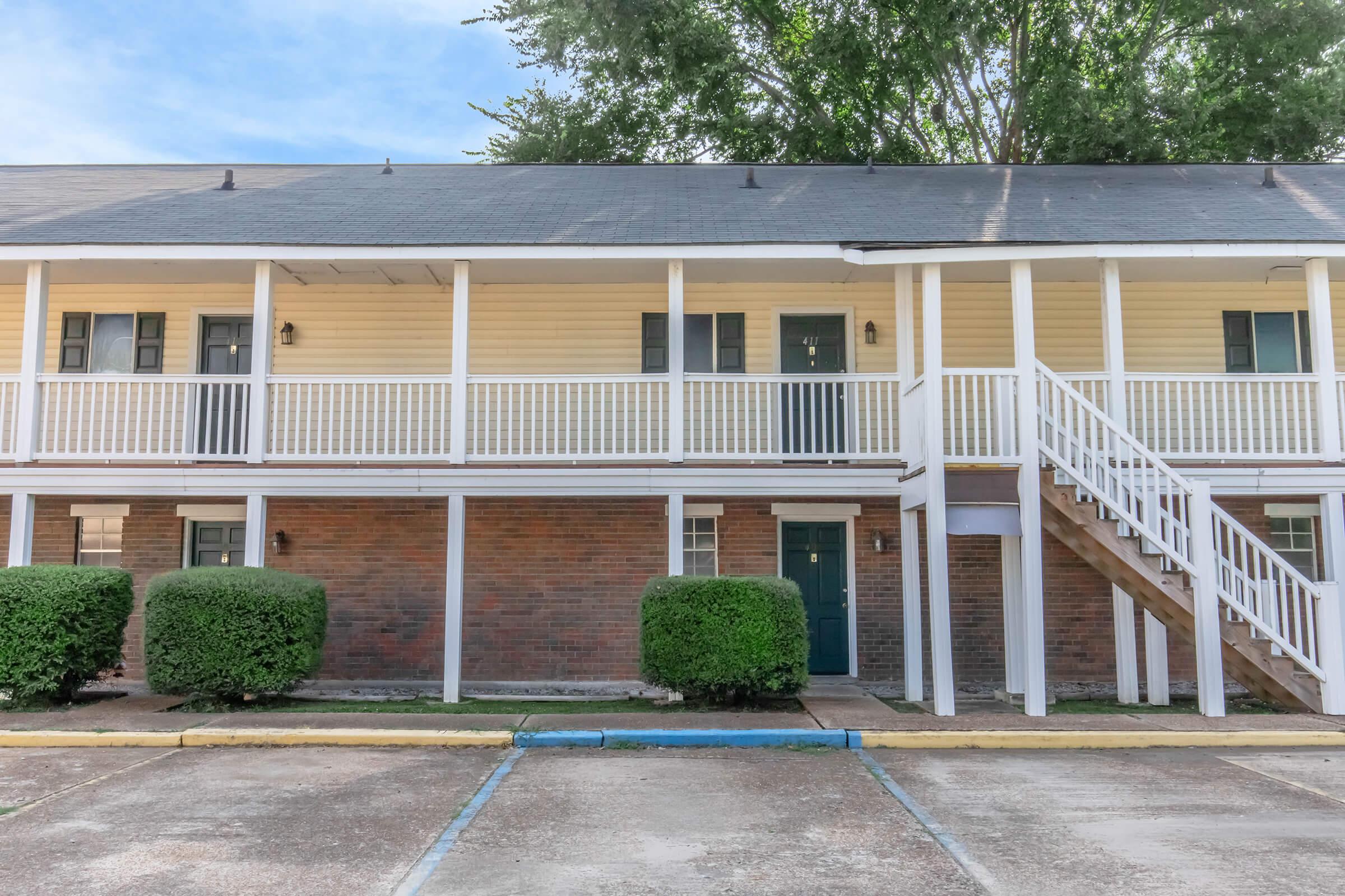 Two-story building with a yellow and brick exterior. The upper level features white railings with several doors. The lower level has a staircase leading up and is flanked by neatly trimmed bushes. The parking area is visible in front, with a blue line marking a parking space.