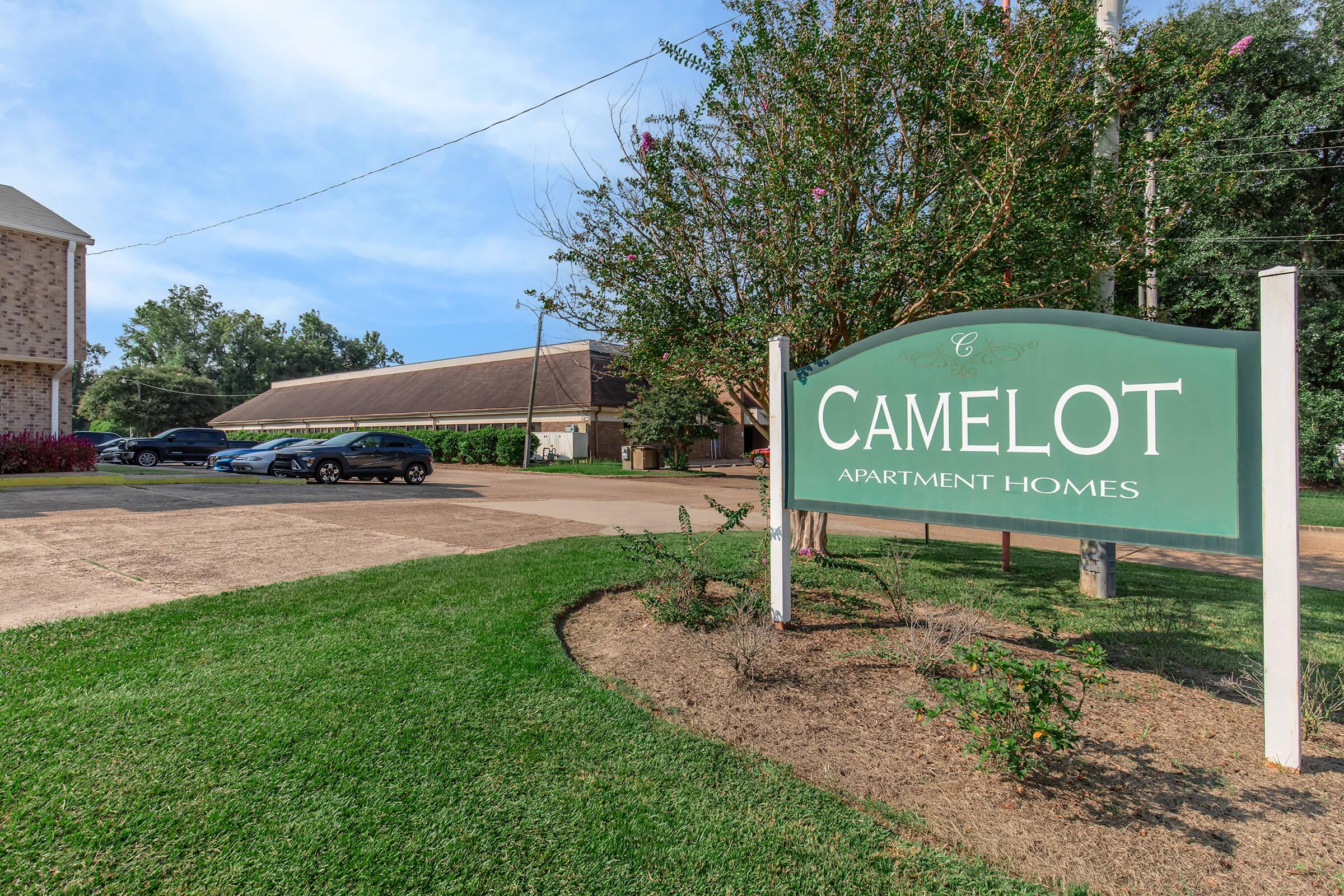 Sign for Camelot Apartment Homes displayed prominently in a landscaped area, with a gravel parking lot and cars parked nearby. The background features trees and a building, all under a clear blue sky.