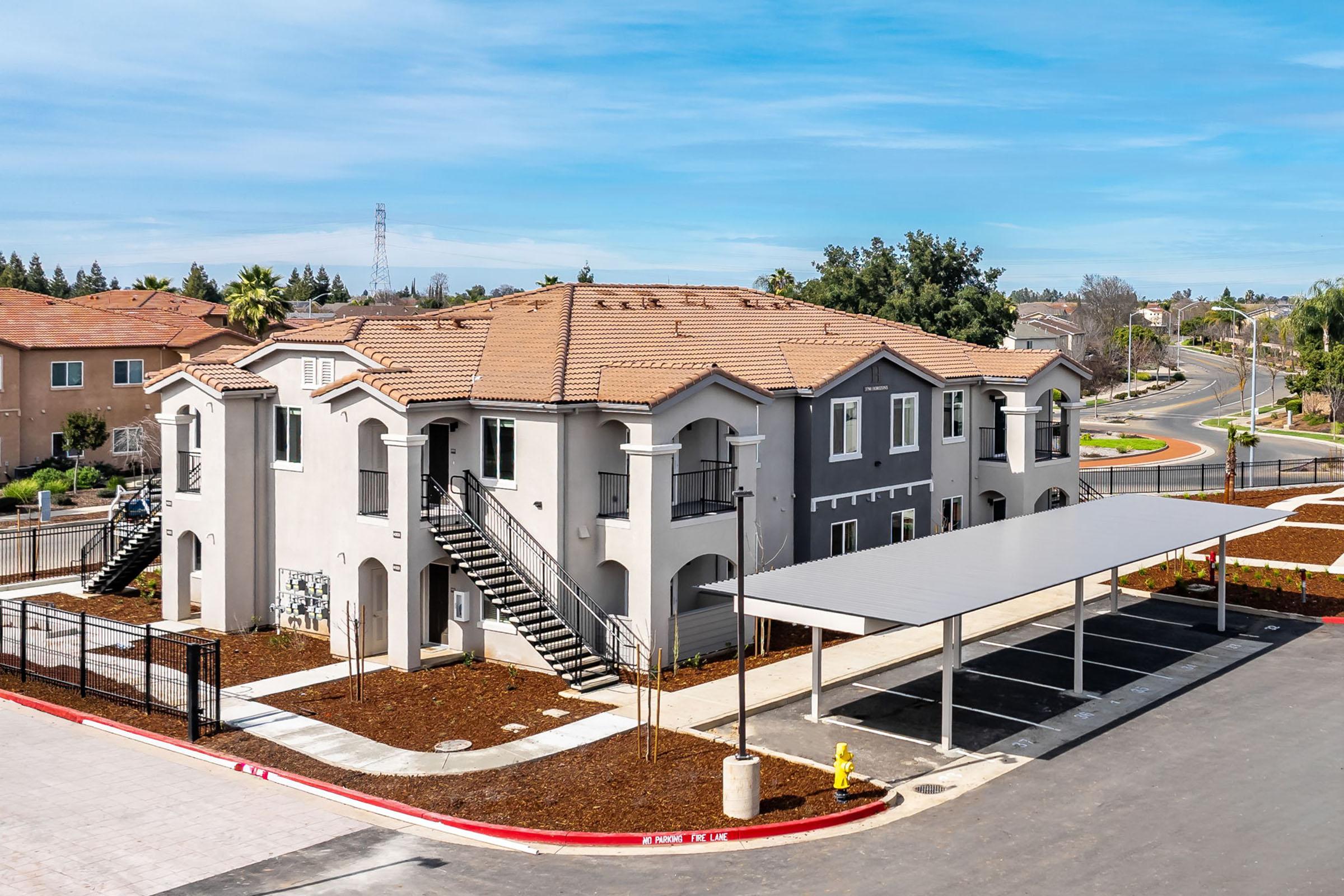 A modern multi-unit residential building with a tiled roof, featuring multiple staircases and a covered parking area. Surrounding landscaping includes freshly laid mulch and a few trees. The scene captures a clear blue sky and nearby roadways.