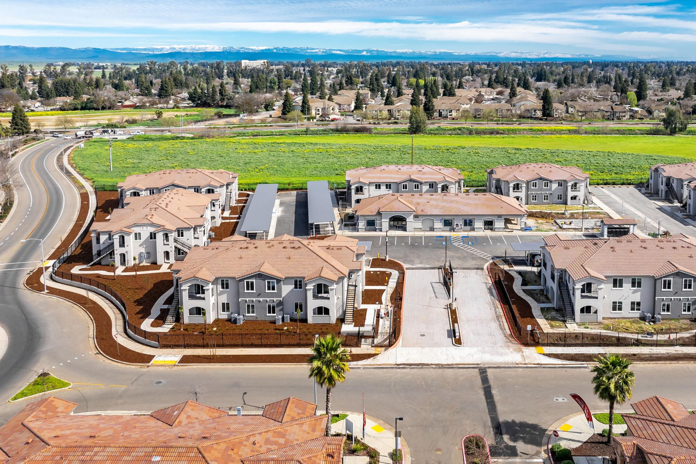 Aerial view of a residential neighborhood featuring modern multi-story buildings with terracotta roofs, surrounded by landscaped grounds. In the background, fields and distant mountains are visible, while a curved road runs alongside the complex. The setting appears peaceful and well-maintained.