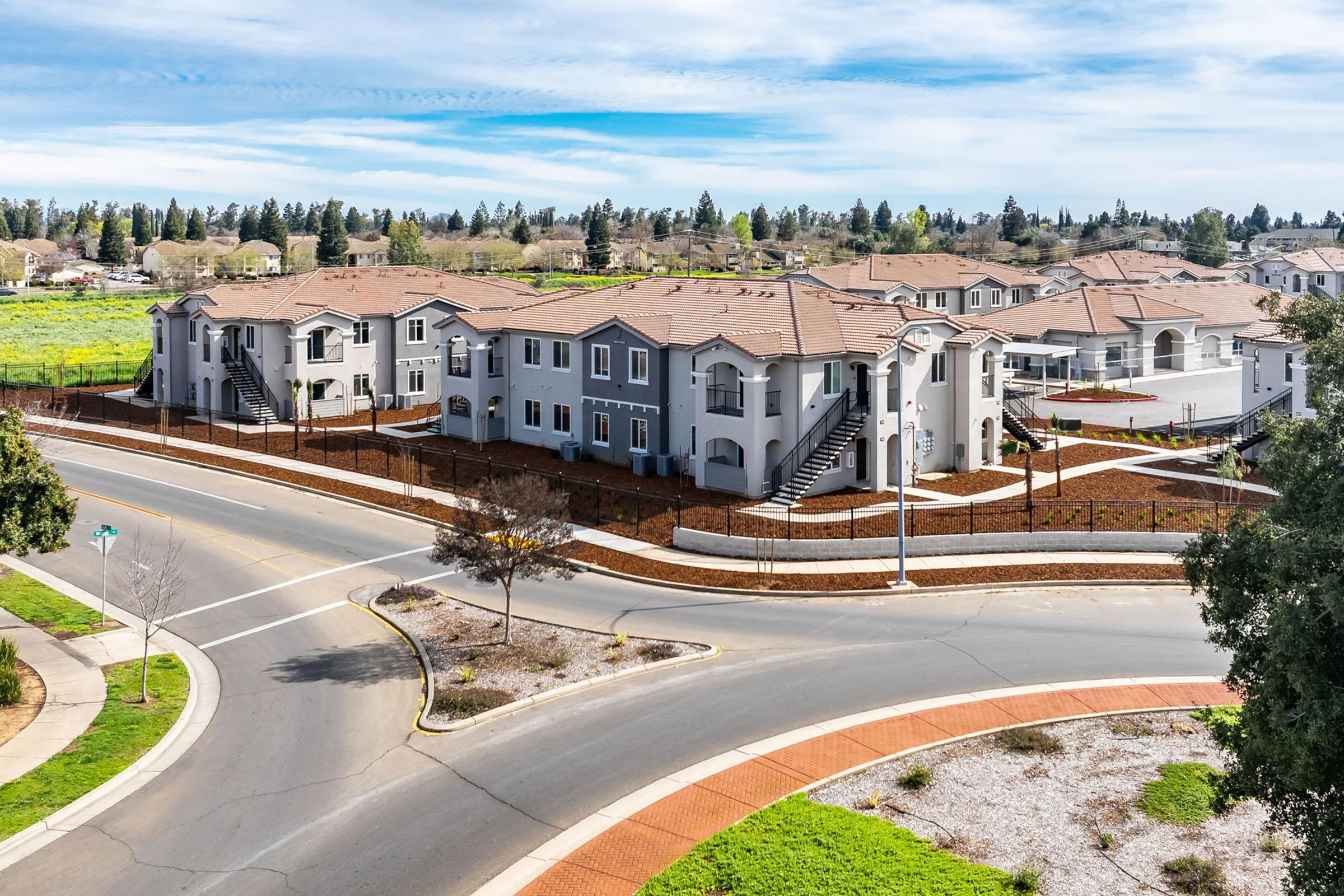 Aerial view of a modern residential complex featuring multiple two-story buildings with beige exteriors and brown roofs. The complex is surrounded by landscaped areas and a circular road, set against a backdrop of greenery and trees under a clear blue sky.
