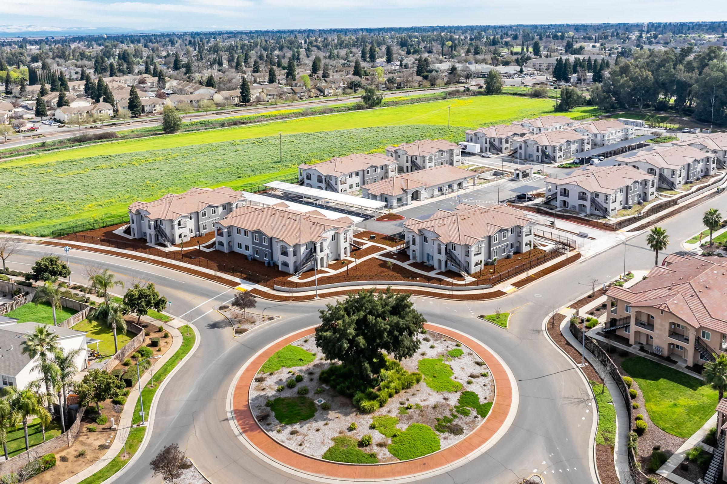 Aerial view of a residential area featuring modern apartment buildings arranged around a circular intersection. The surrounding landscape includes green fields and trees, with well-maintained landscaping in the foreground. Bright, clear sky adds to the tranquil atmosphere of the neighborhood.