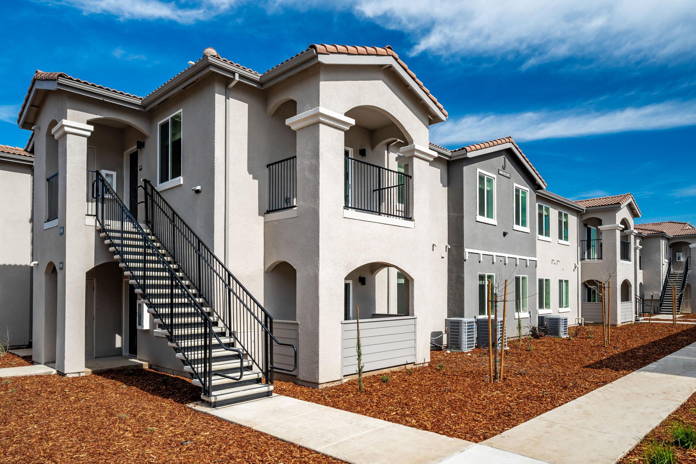 Two-story apartment complex with a modern design featuring arched doorways and balconies. The exterior is painted in light colors, surrounded by landscaped areas with mulch and small trees. A metal staircase leads to the upper level, with sky and clouds visible in the background.