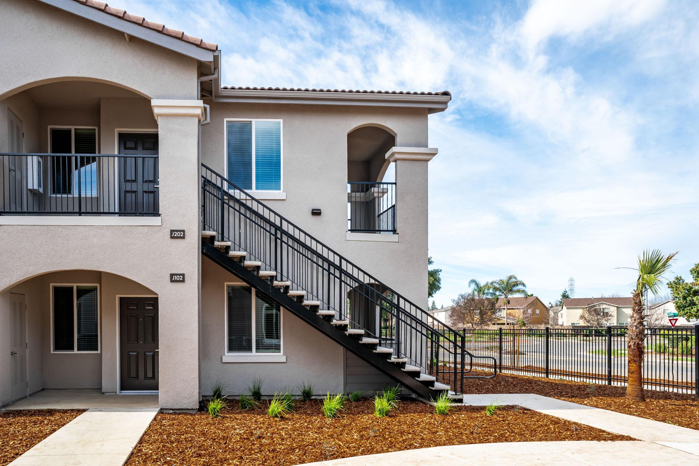 A modern two-story apartment building featuring a staircase leading to the second floor. The exterior is painted in light gray with dark trim, and there's a landscaped area with small plants and mulch. In the background, trees and a clear blue sky are visible.