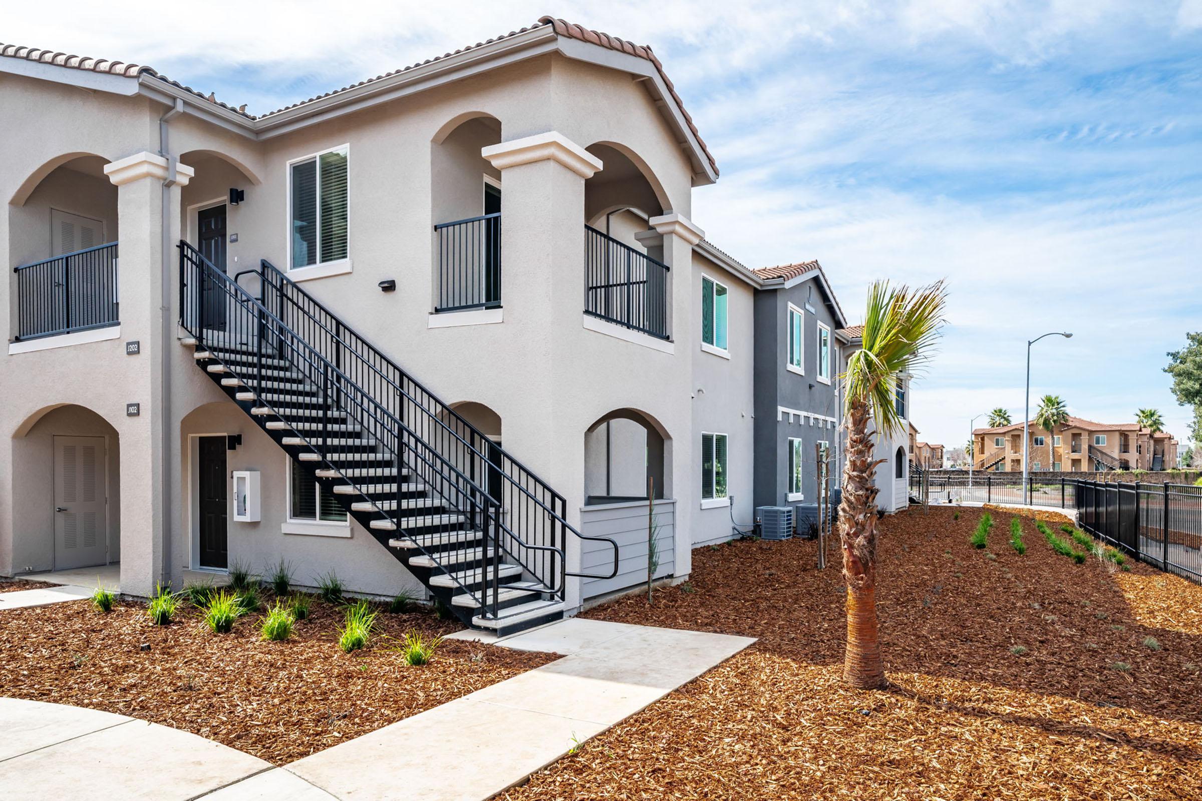 Exterior view of a two-story residential building featuring a staircase on the left side. The building has a modern design with balconies, surrounded by landscaped areas with mulch and small plants. In the background, there are other homes and palm trees under a clear blue sky.