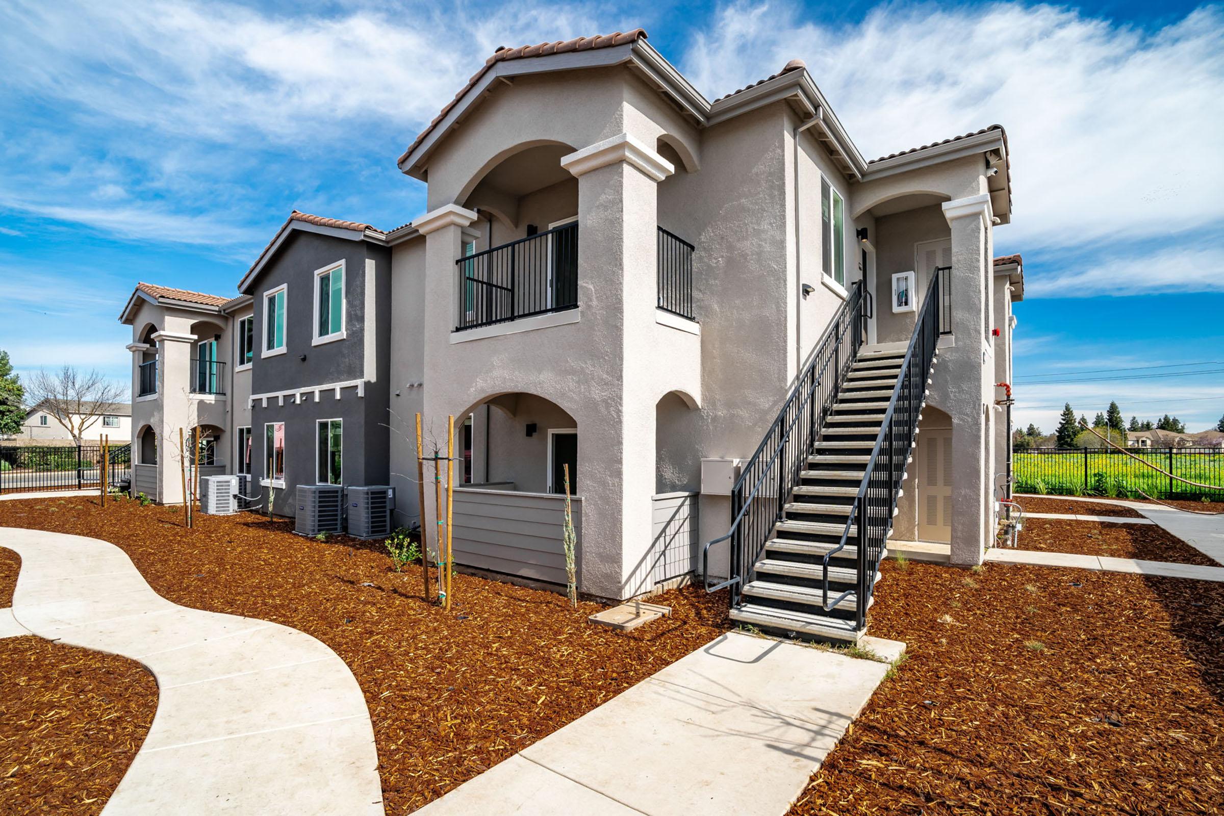 Two-story apartment building with a modern design, featuring light gray exterior, arched entrances, and black railings. A curved pathway lined with mulch leads to the building, and lush greenery is visible in the background under a bright blue sky with scattered clouds.