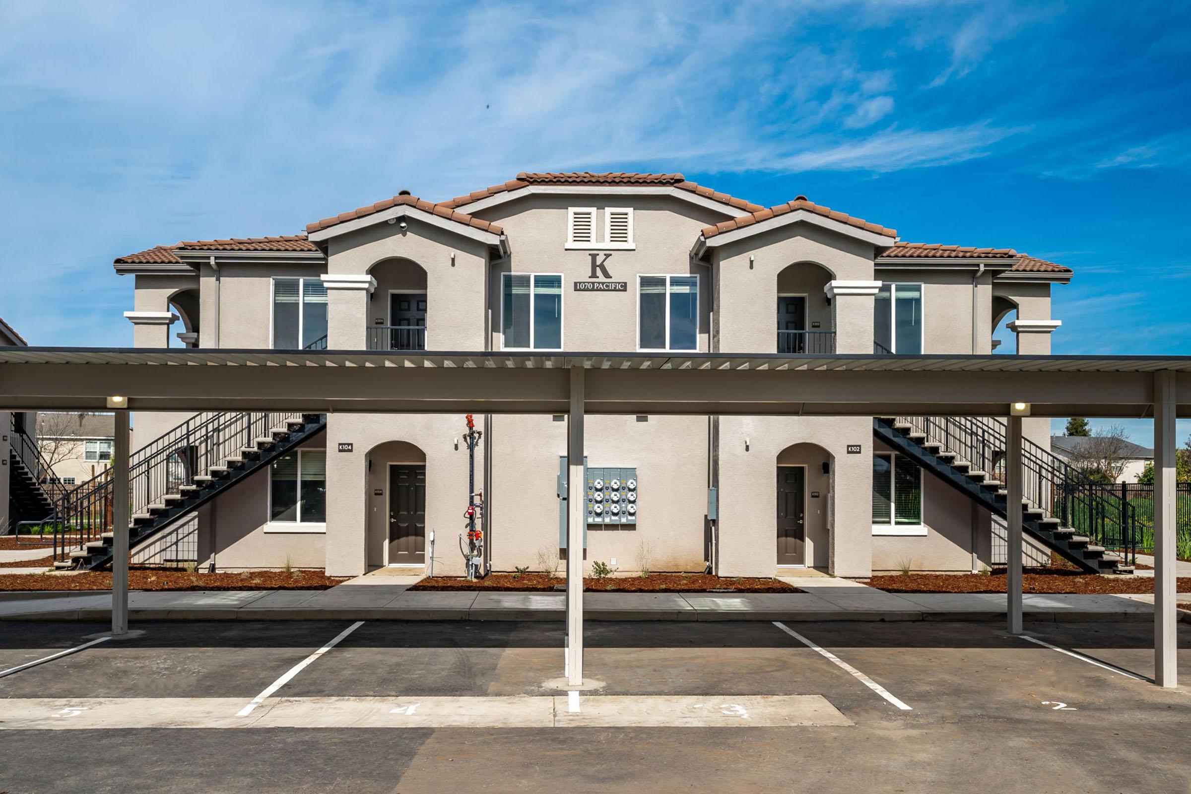 Two-story apartment building with a stucco exterior, featuring staircases on either side leading to balconies. The building has multiple doors and mailboxes at the front. A covered parking area is in the foreground with empty parking spaces. Clear blue sky with few clouds in the background.