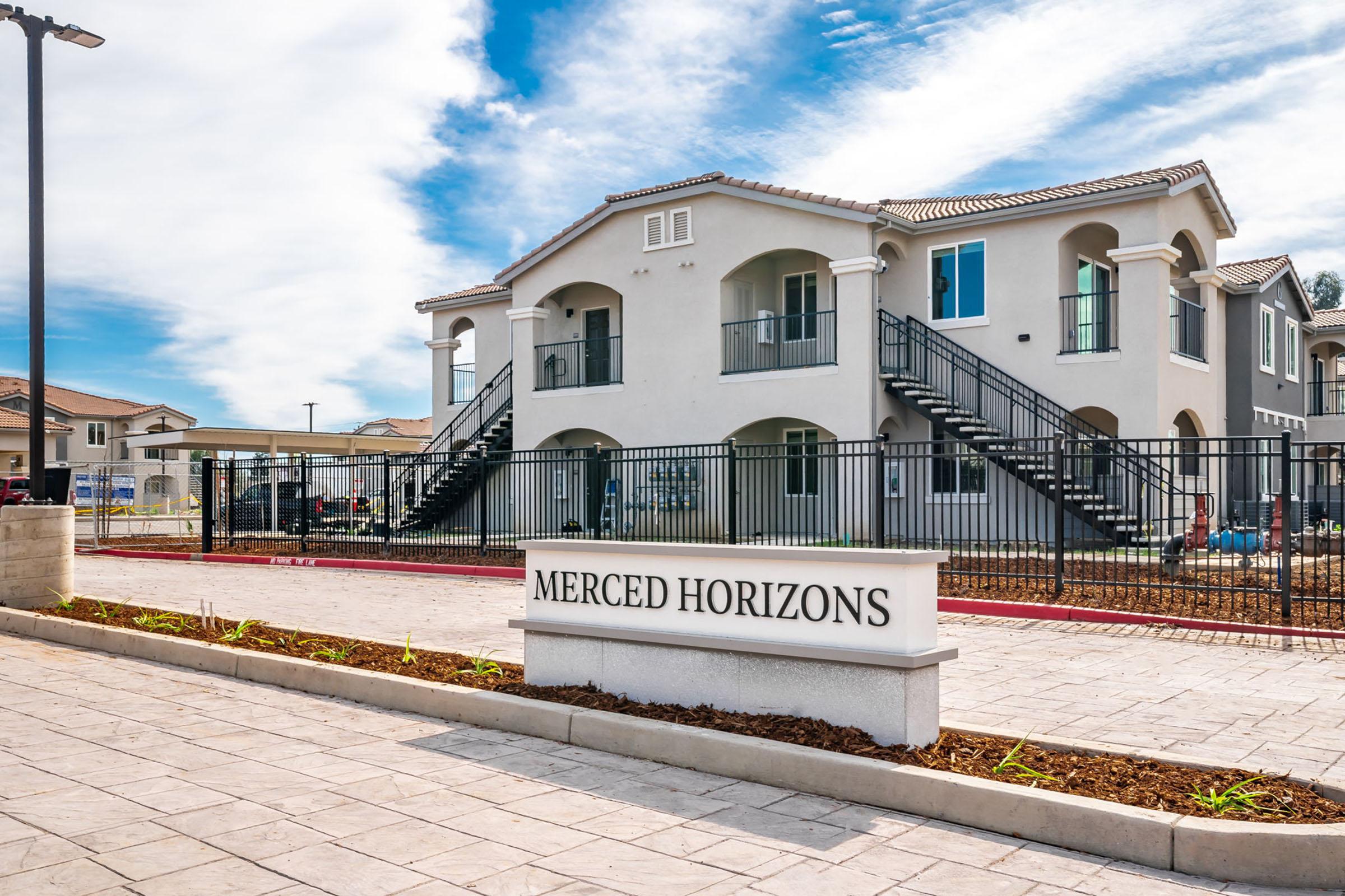 A modern apartment complex named "Merced Horizons" featuring a two-story building with a light-colored exterior, balconies, and staircases. The entrance is marked by a prominent sign, and the surrounding area has neatly landscaped pavement. The sky is partly cloudy, adding to the bright atmosphere.