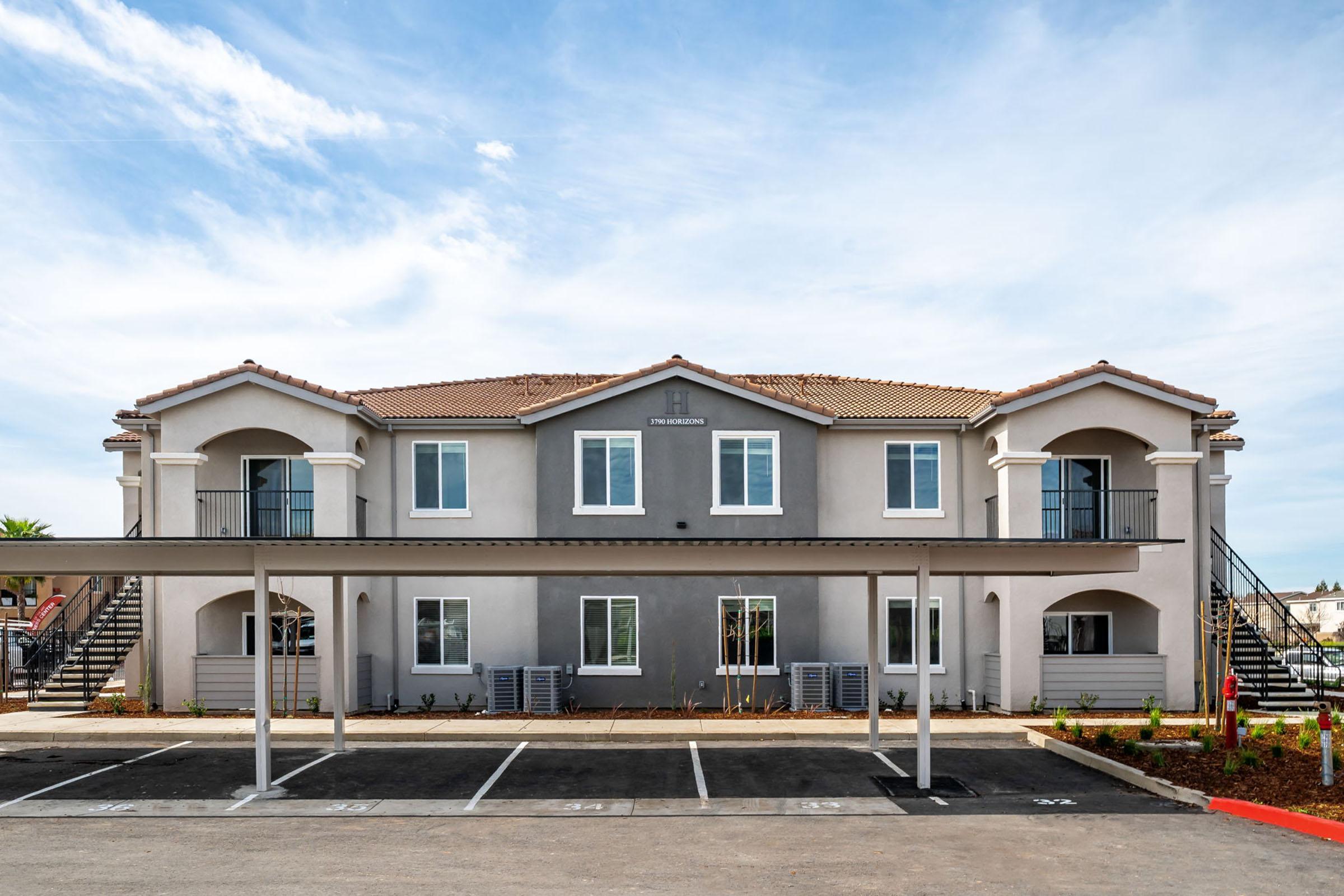 A multi-unit residential building with a modern design, featuring a gray exterior and a tile roof. It has two levels with balconies and a staircase. The foreground shows designated parking spaces under a canopy. There are landscaped areas with plants along the building's sides. The sky is clear with some clouds.
