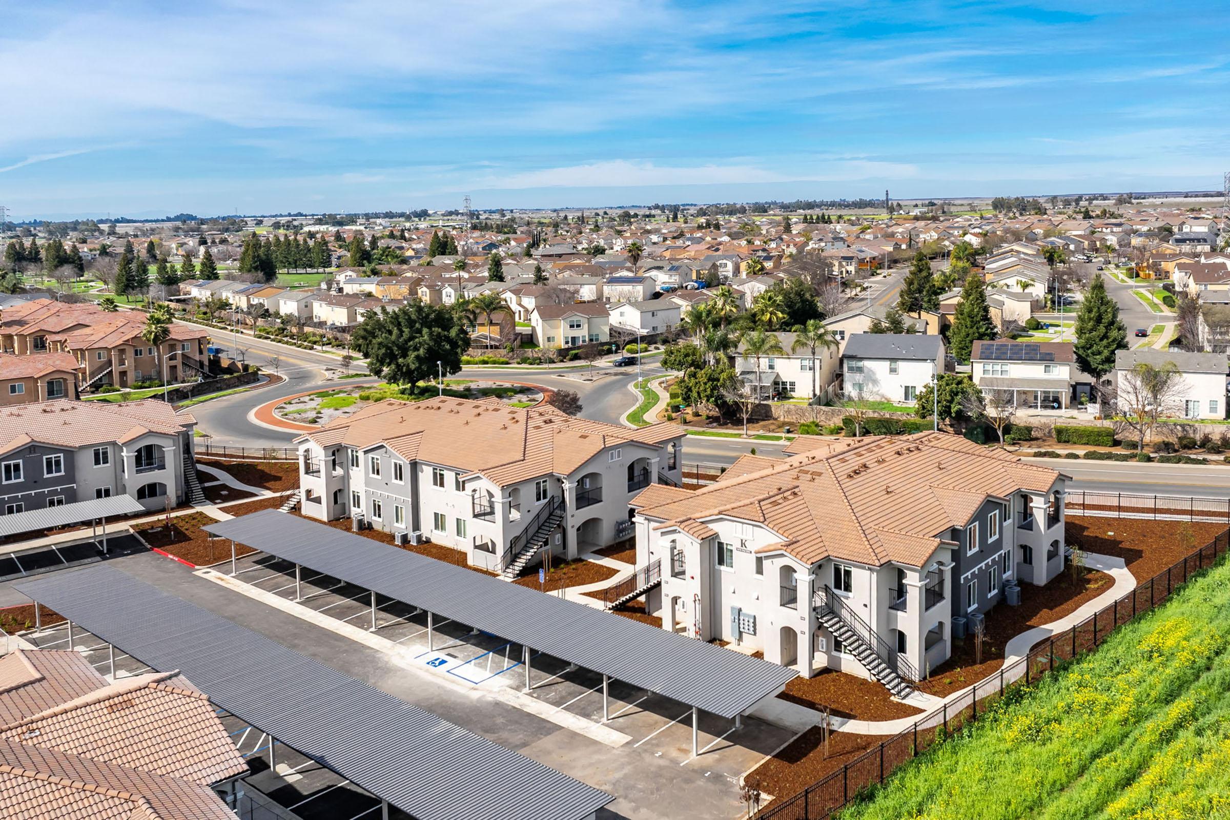 Aerial view of a residential neighborhood featuring modern multi-family buildings with terracotta roofs, surrounded by green landscaping and a parking area with shaded carports. The scene includes streets, houses, and open spaces under a clear blue sky.