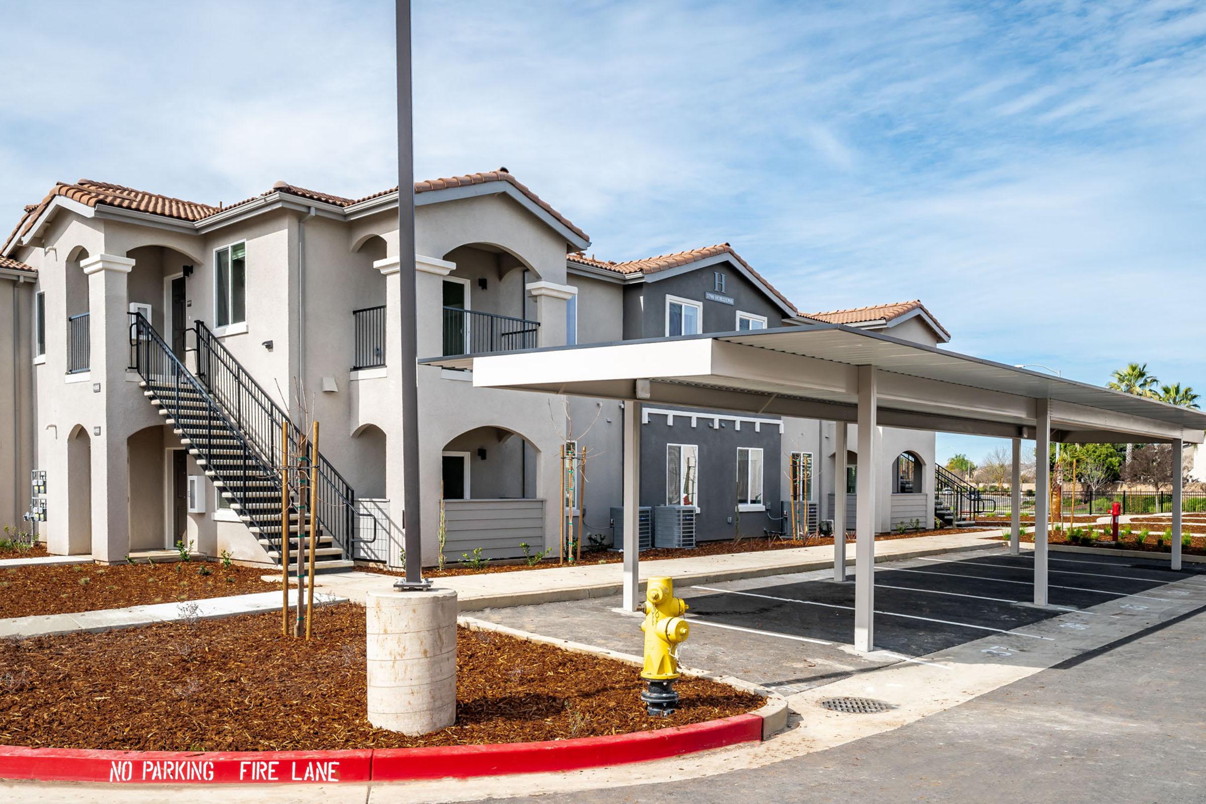 A modern two-story apartment building with a stairway, surrounded by newly landscaped grounds. Covered parking spaces are visible in front, with a fire hydrant nearby. A sign indicates no parking in the fire lane, and the sky is clear with a few clouds.