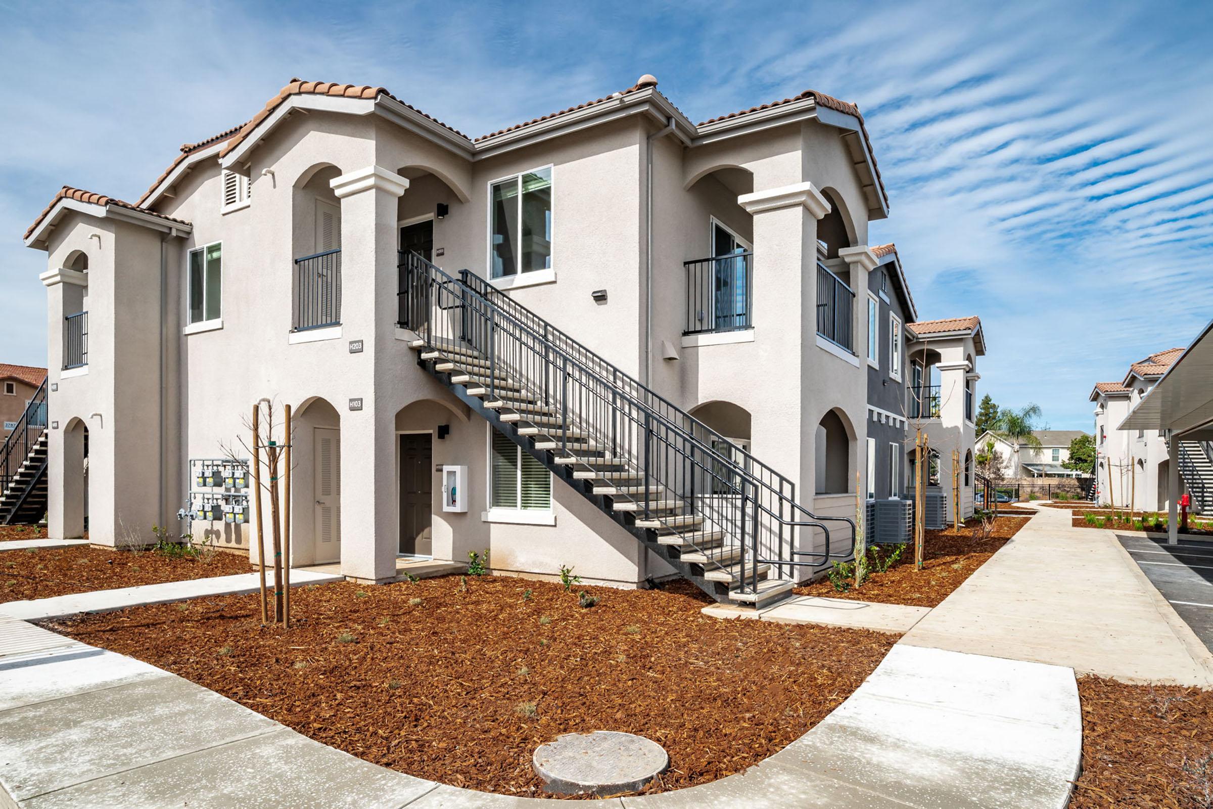 Exterior view of a multi-unit residential building with two stories. The structure features a landscaped yard with mulch, a sidewalk, and a staircase leading to upper units. A clear blue sky is visible, enhancing the bright, inviting atmosphere of the property.