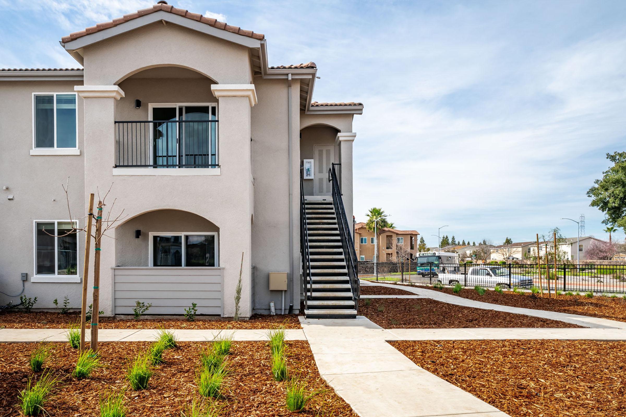 A modern two-story apartment building with a beige exterior, featuring balconies and a staircase. The surrounding landscape includes freshly mulched areas and small plants, with a paved walkway leading to the entrance. In the background, there are additional buildings and a clear blue sky.