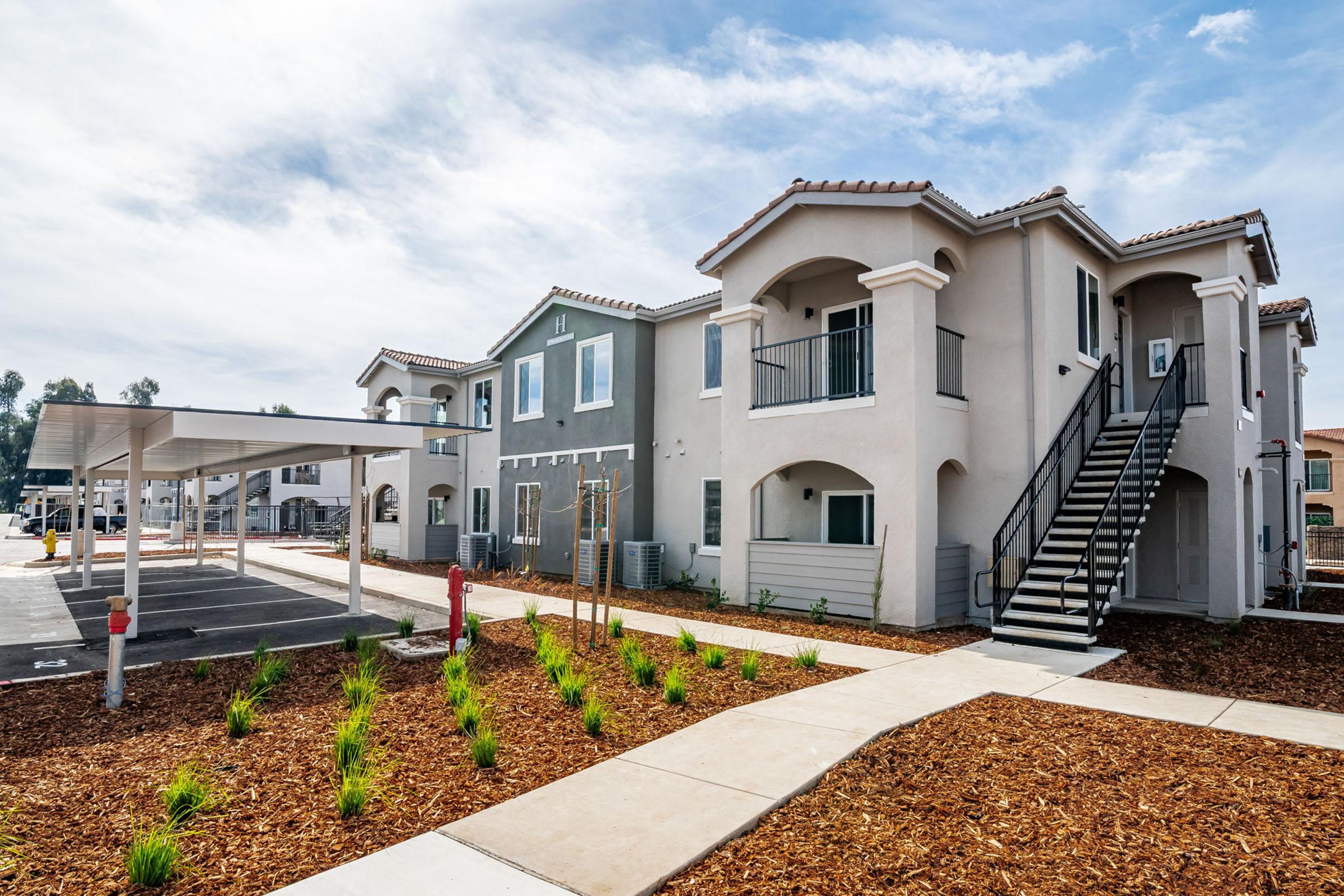 Modern two-story apartment complex with a combination of light and dark gray exteriors. Features include balconies, a staircase, and a landscaped area with mulch and small plants. The building is set against a partly cloudy sky, with a parking area and covered spaces in the foreground.