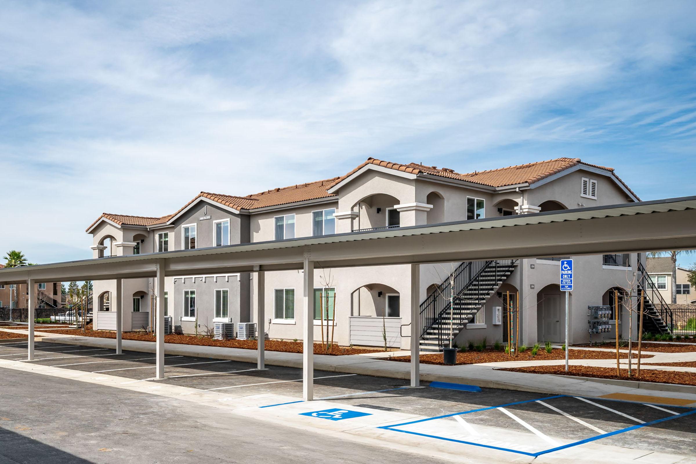 A modern two-story apartment building with a stucco exterior and a red-tiled roof. The structure features multiple windows and outdoor staircases. In the foreground, there are parking spaces, including designated spots for accessibility, and a covered parking area. The sky is clear with a few clouds.