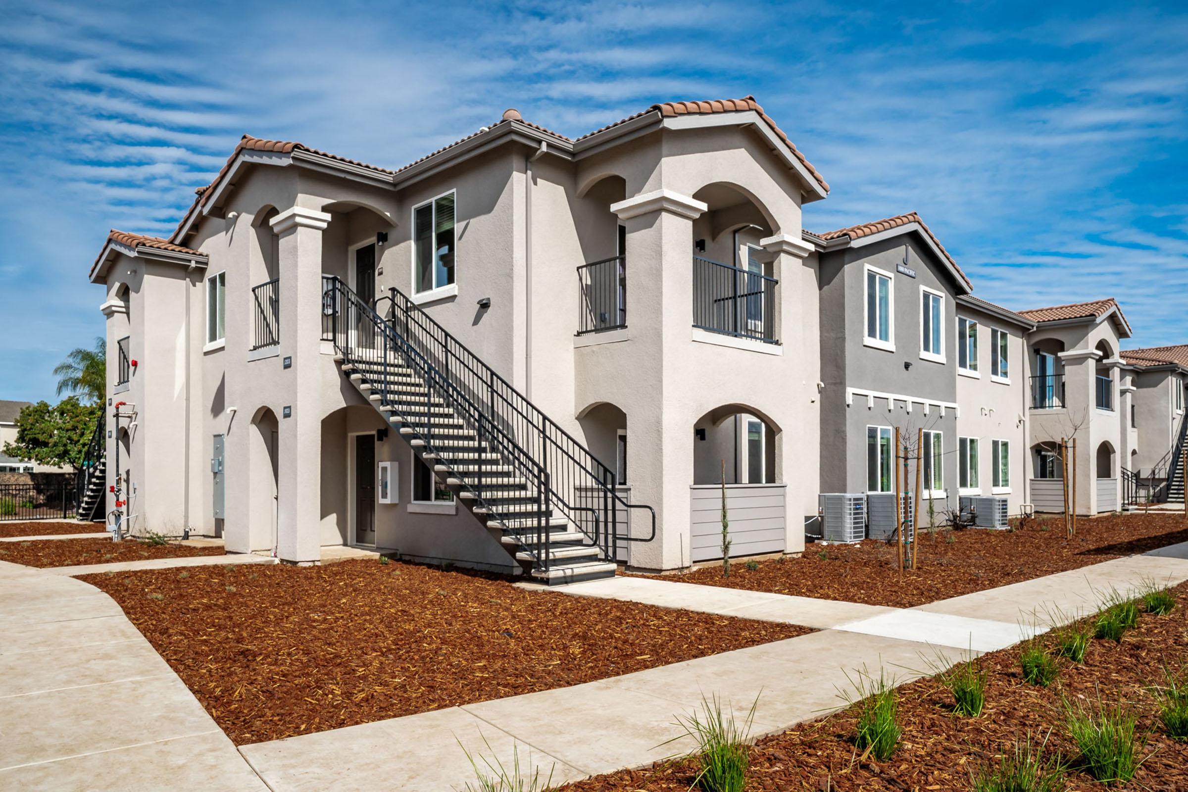 Modern two-story apartment building with a light-colored exterior, featuring balconies and stairs. The surrounding area has freshly landscaped mulch and small plants, under a bright blue sky with scattered clouds.