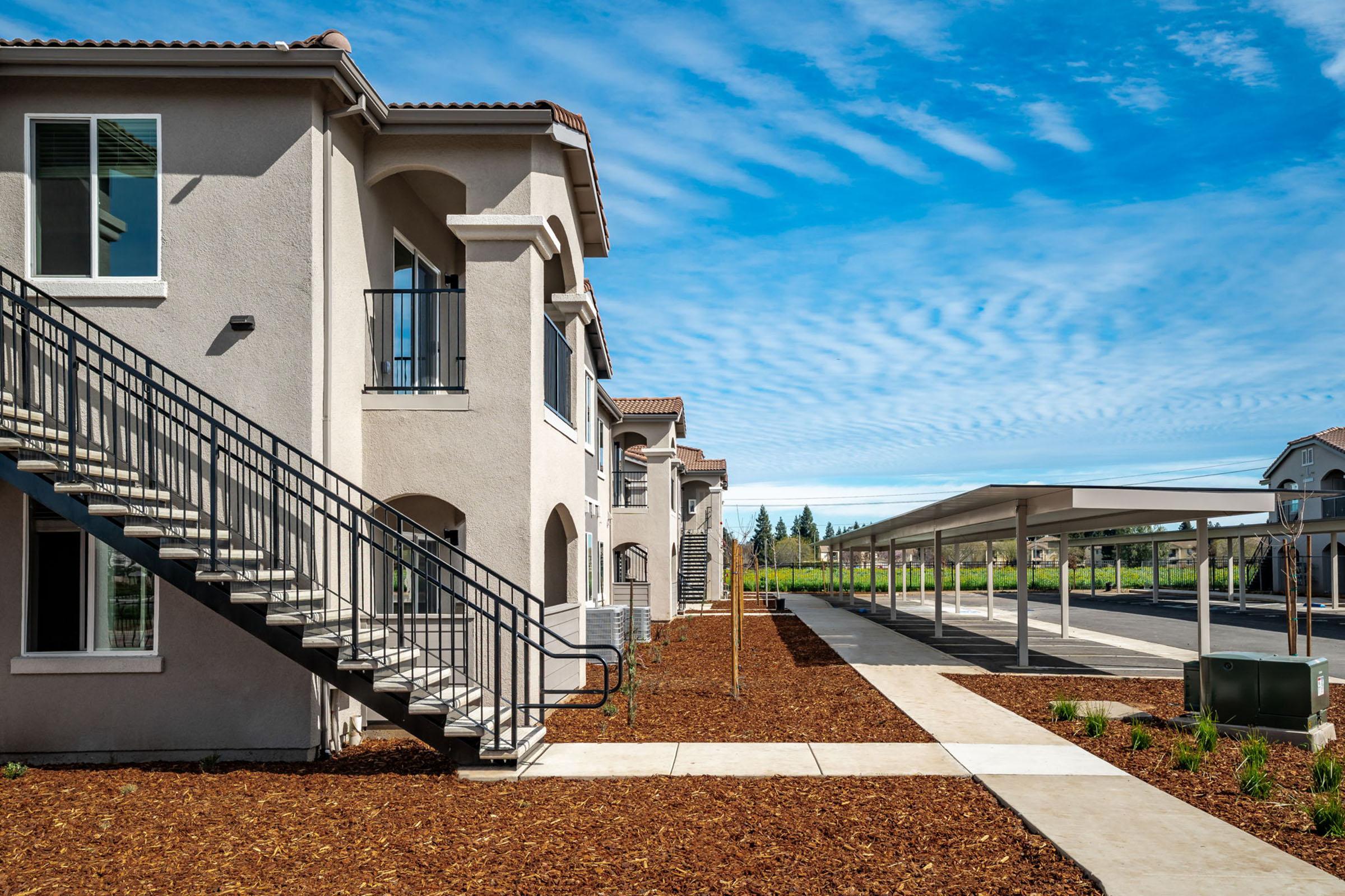 A view of a residential complex featuring beige multi-story buildings with metal stairs, surrounded by landscaped grounds and a covered parking area, under a blue sky with scattered clouds. Paths of concrete lead through the landscaping, creating a welcoming outdoor space.