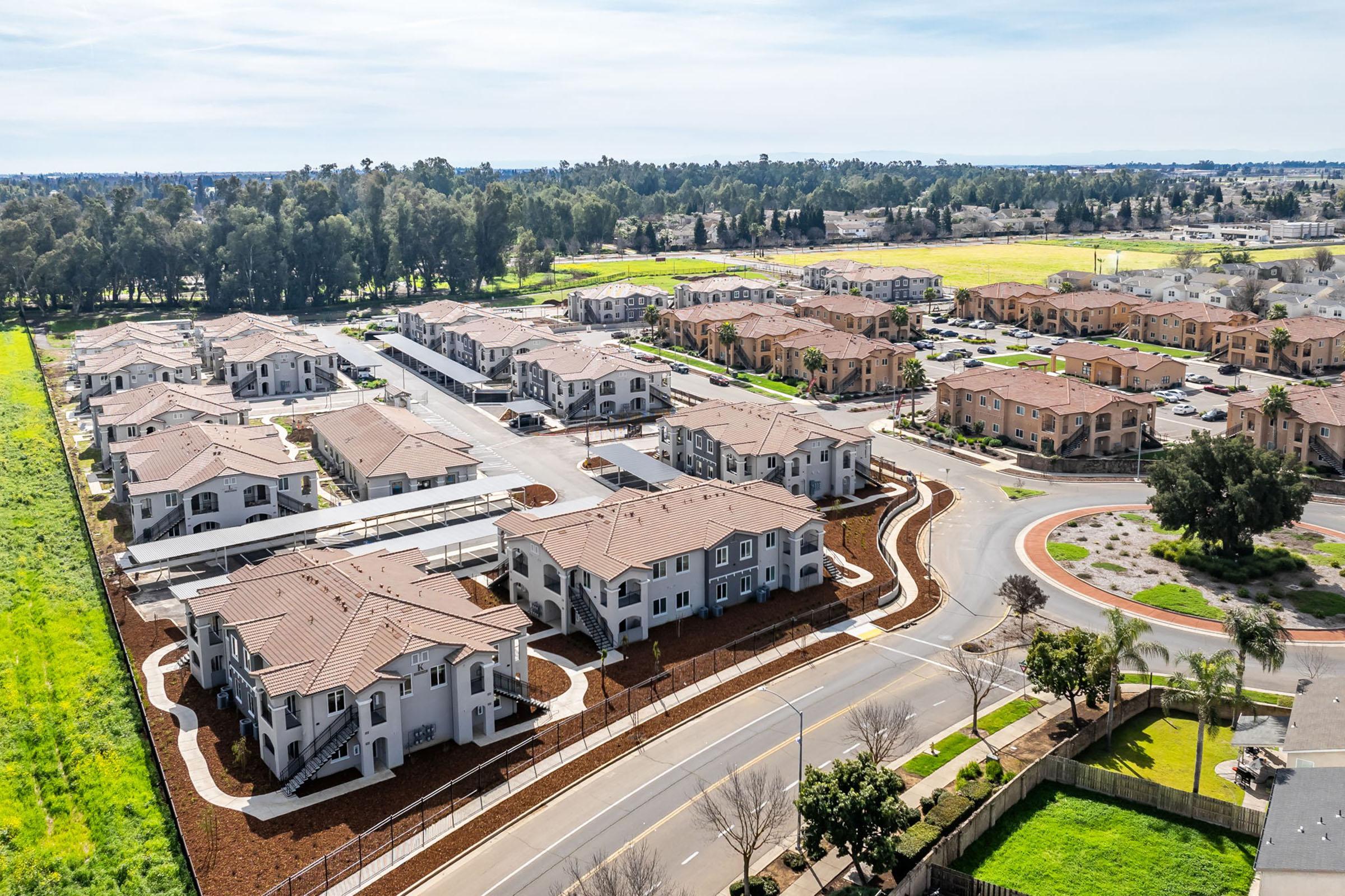 Aerial view of a residential development featuring multiple apartment buildings arranged around circular and straight roadways. Lush green spaces and trees are visible in the background, with additional homes or buildings further in the distance. The scene captures a blend of urban living and natural surroundings.