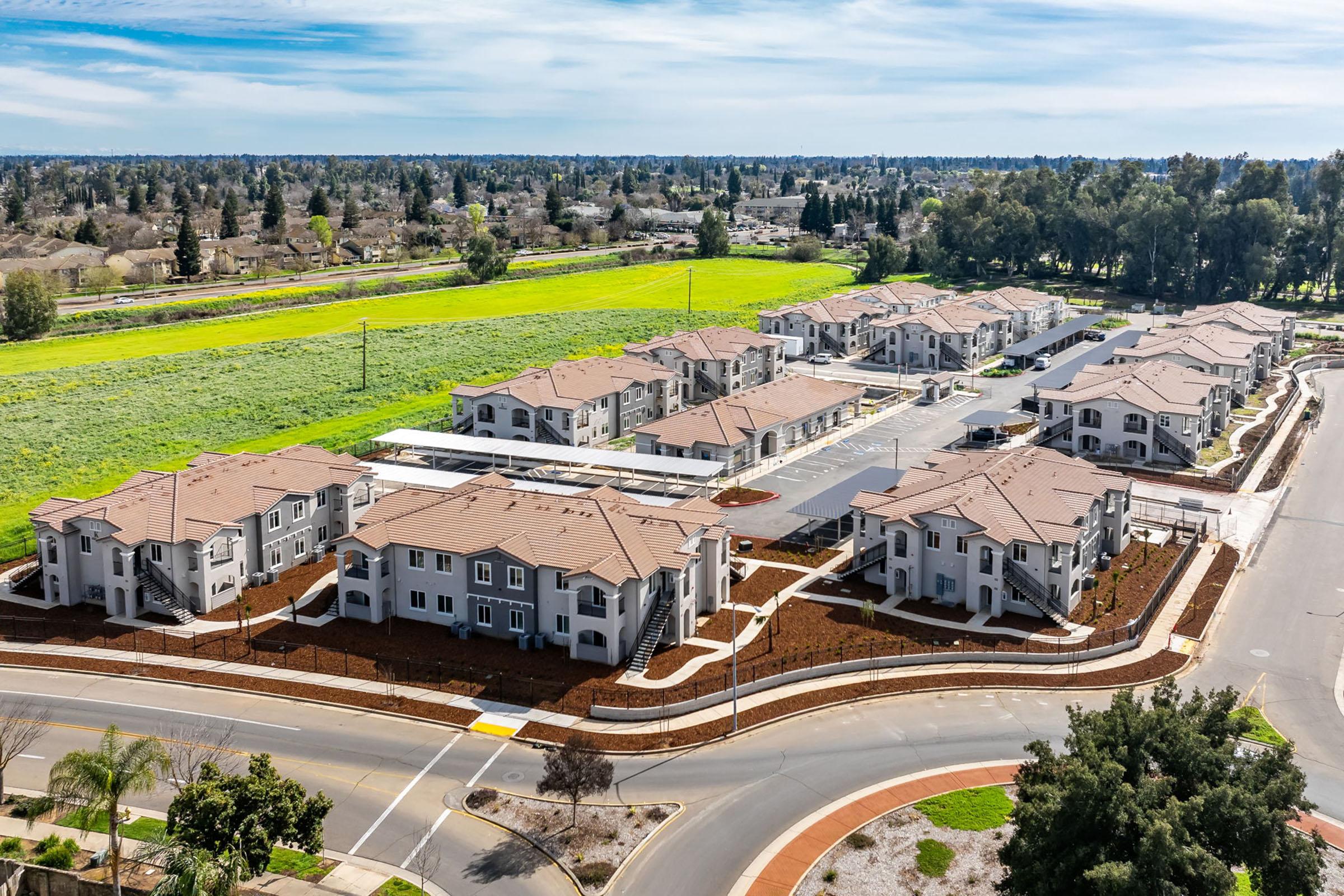 Aerial view of a modern residential complex featuring multiple two-story buildings with brown roofs, surrounded by landscaped areas. In the background, a lush green field and trees are visible, along with a curved road leading into the complex.