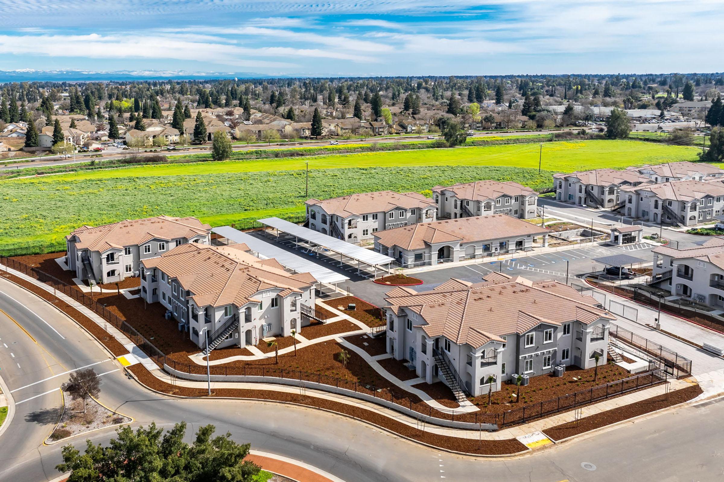 Aerial view of a residential complex featuring multiple buildings with brown roofs and landscaped grounds. Surrounding the complex are green fields and suburban neighborhoods under a clear blue sky. Roads and parking areas are visible in the foreground, creating a structured layout.