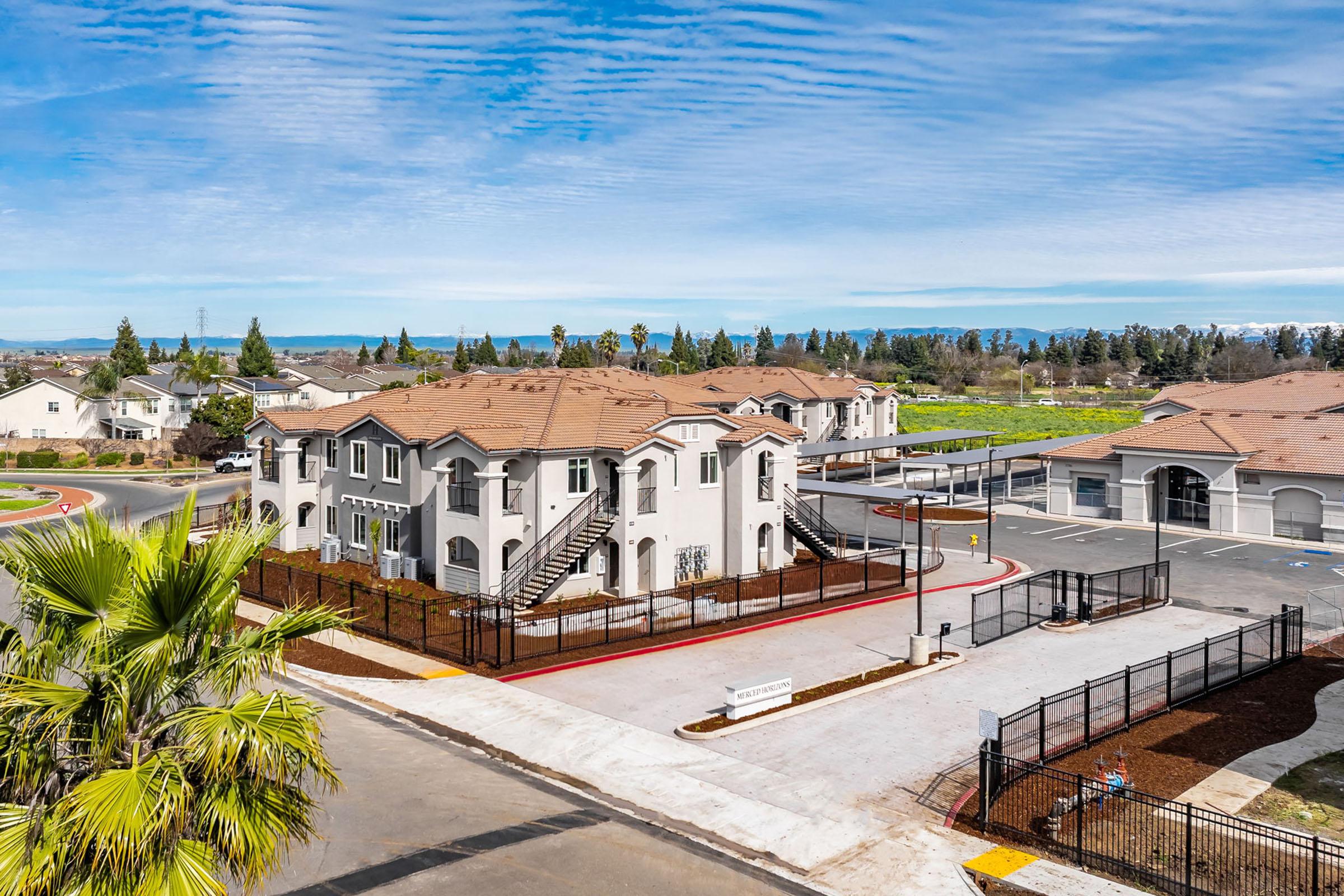 Aerial view of a modern residential complex with beige buildings featuring staircases. Surrounding the complex are landscaped areas, palm trees, and a clear blue sky. In the background, rolling hills and vegetation can be seen, along with a paved parking area. The setting appears peaceful and well-maintained.