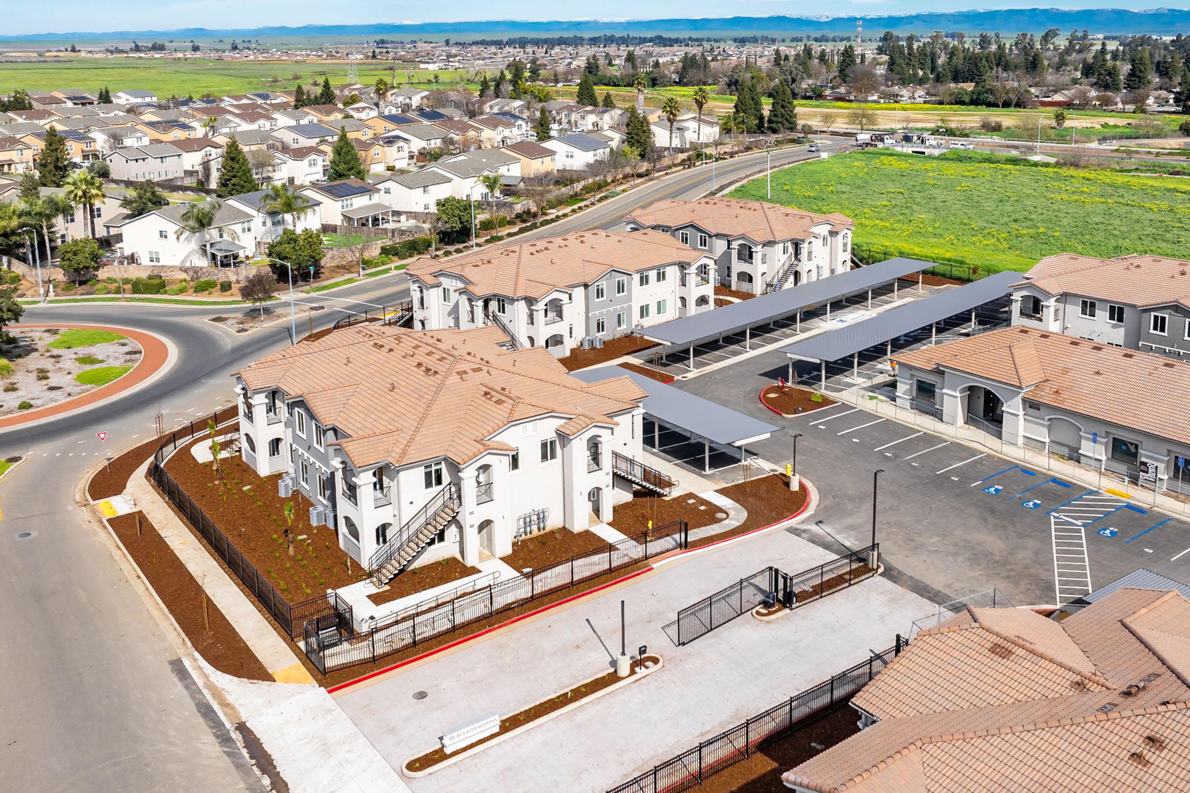 Aerial view of a residential complex featuring multiple two-story buildings with tan roofs, surrounded by landscaped areas and carports. Nearby, suburban houses and green fields are visible, along with a circular road. The scene is bright and shows a well-maintained community environment.