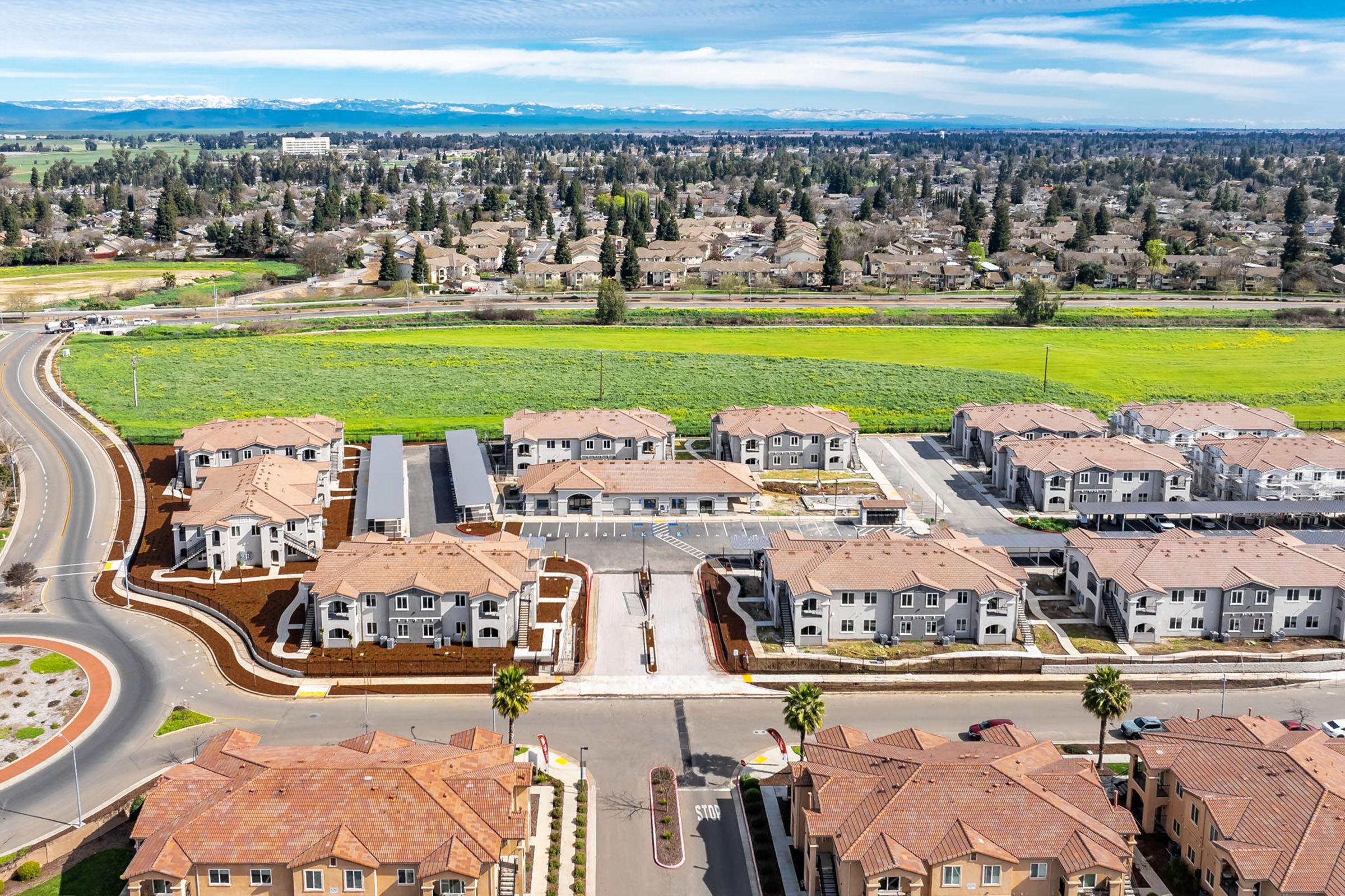 Aerial view of a suburban residential area featuring multiple apartment buildings with tiled roofs, landscaped grounds, and parking areas. In the background, lush green fields and hills can be seen, with a clear blue sky above. The layout includes roadways and various residential structures.