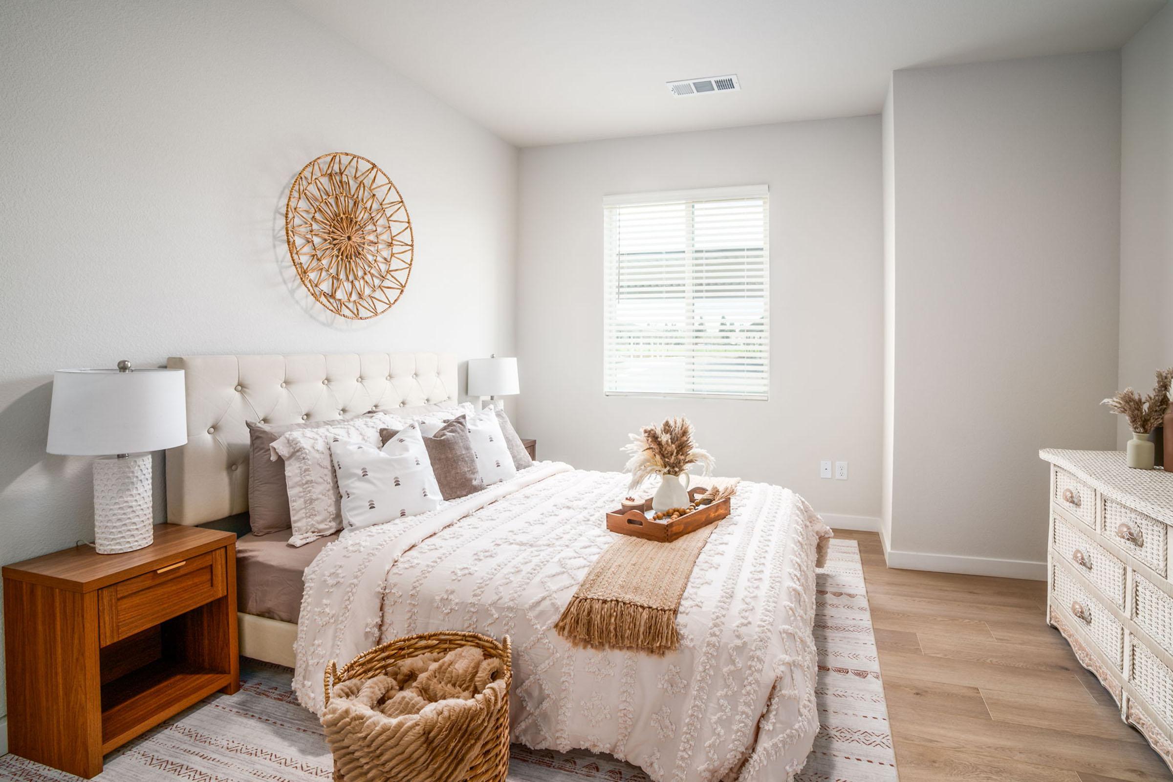 A cozy, modern bedroom featuring a queen-sized bed with decorative pillows and a soft throw blanket. There's a woven wall hanging above the headboard, two bedside lamps on wooden nightstands, and a dresser. Natural light filters in through a window, illuminating the neutral color palette and warm wooden floors.