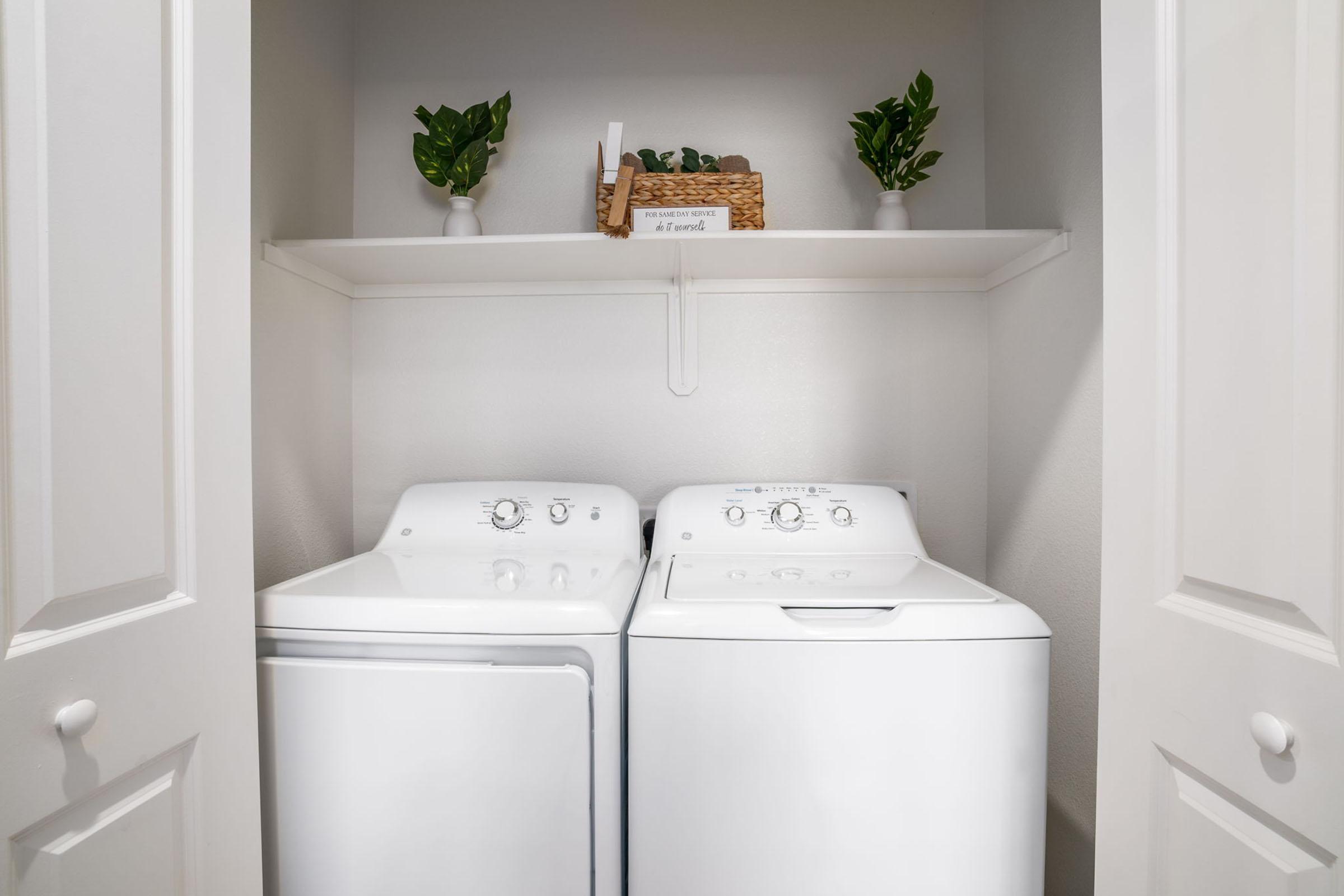 A clean laundry room featuring a washing machine and dryer side by side. Above them is a shelf with two small potted plants and a wicker basket, all set against a light-colored wall inside a closet with white doors.