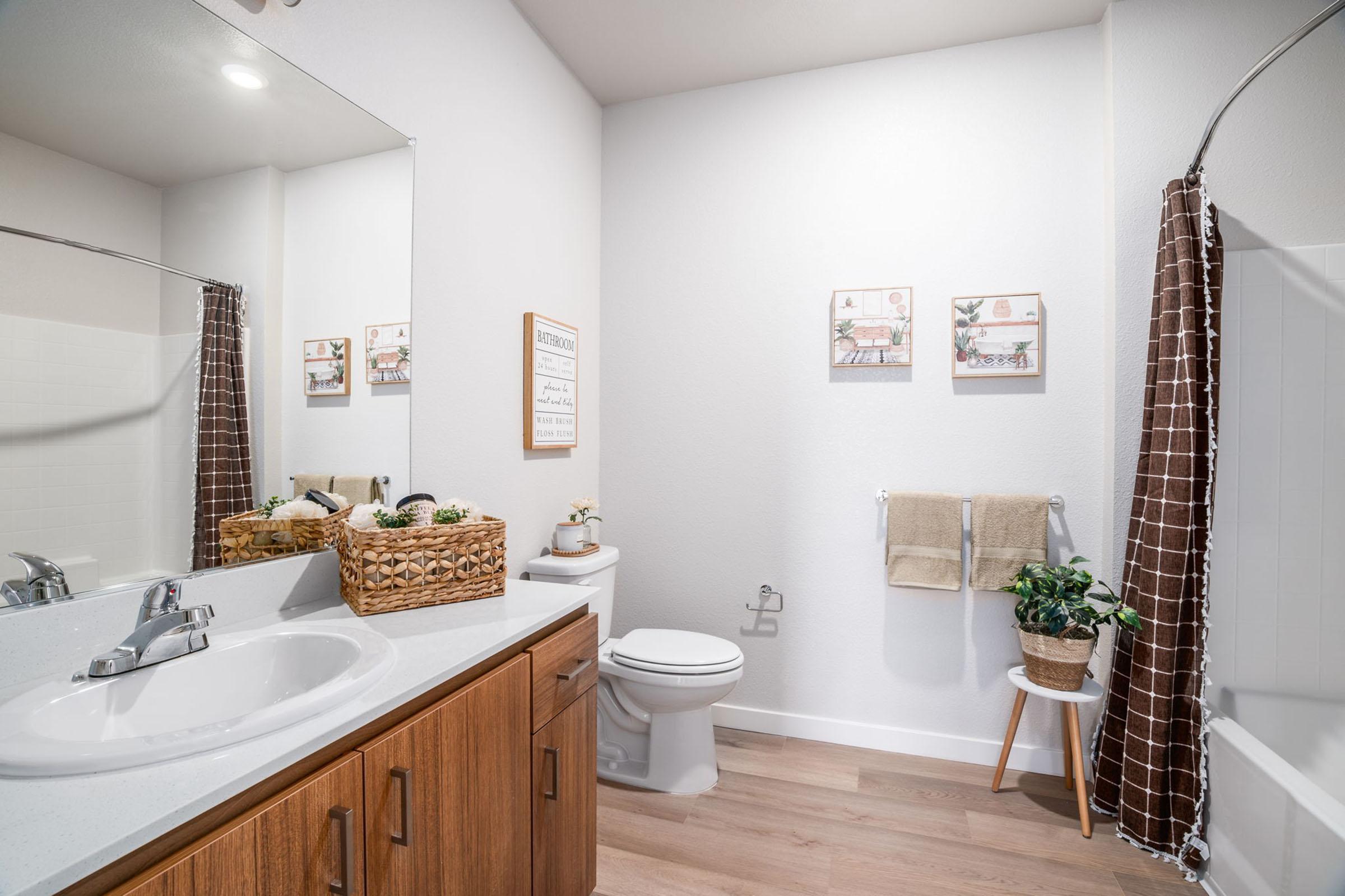 A modern bathroom featuring a double sink vanity with a large mirror, a wooden cabinet below, a toilet, and a bathtub. The décor includes two framed prints on the wall, a towel rack with towels, a wicker basket holding toiletries, and a potted plant on a stool, providing a clean and inviting atmosphere.