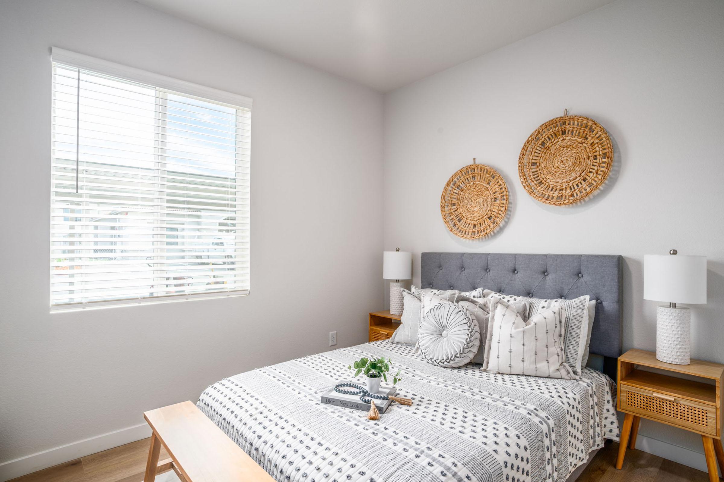 A cozy bedroom featuring a grey upholstered bed with patterned pillows and a cozy throw. Two woven wall hangings are displayed above the bed. There are wooden nightstands with white table lamps on either side, and a window providing natural light, enhancing the room's airy atmosphere.