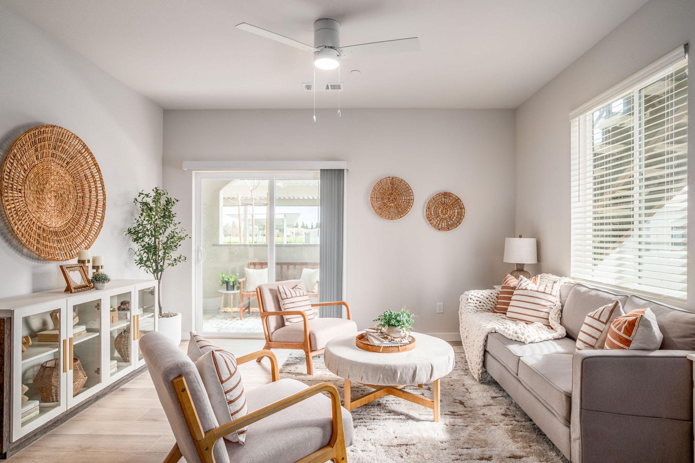 A modern living room featuring a light color palette. It includes a gray sofa, two armchairs, a round coffee table, and decorative wall baskets. There’s a plant and decorative items on display. A ceiling fan is overhead, and a bright window leads to an outdoor space. The cozy atmosphere is enhanced by soft textures and natural light.