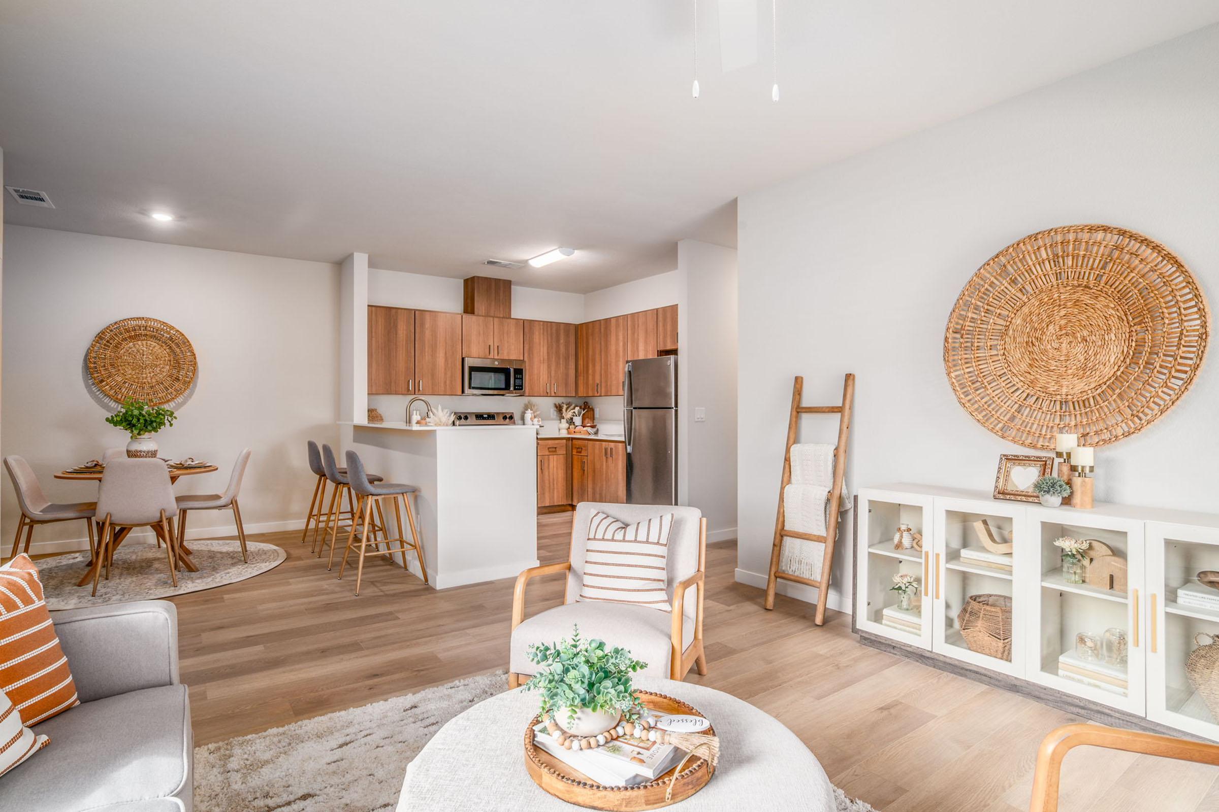 Modern living room and dining area featuring light wood flooring, stylish furnishings, and a neutral color palette. The space includes a cozy seating arrangement with a round coffee table, accent chairs, a dining table for four, and a kitchen area with stainless steel appliances. Decorative wall baskets add warmth.
